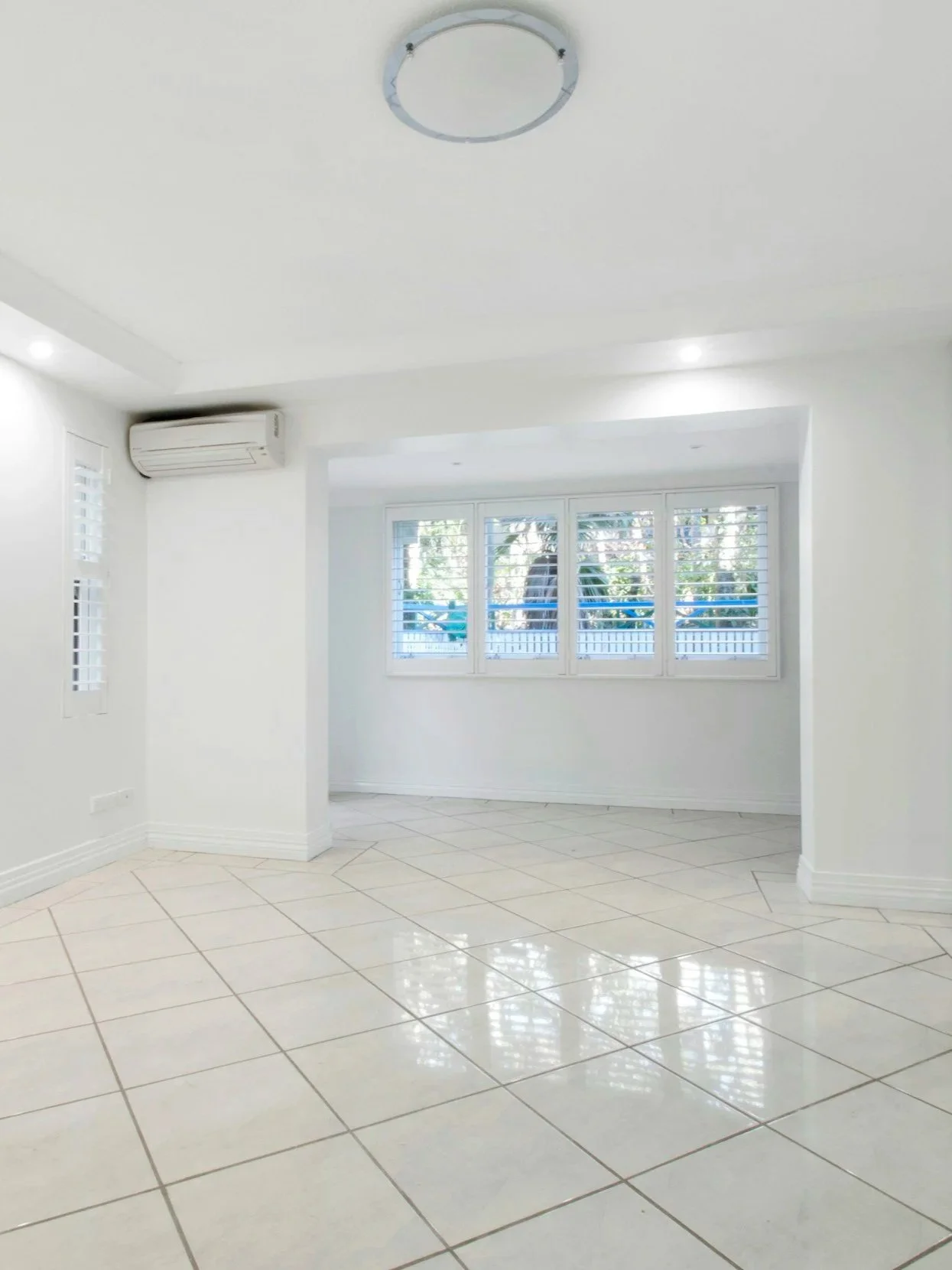Clean, bright living room with white walls, large windows with blinds, tiled floor, and ceiling light fixture.