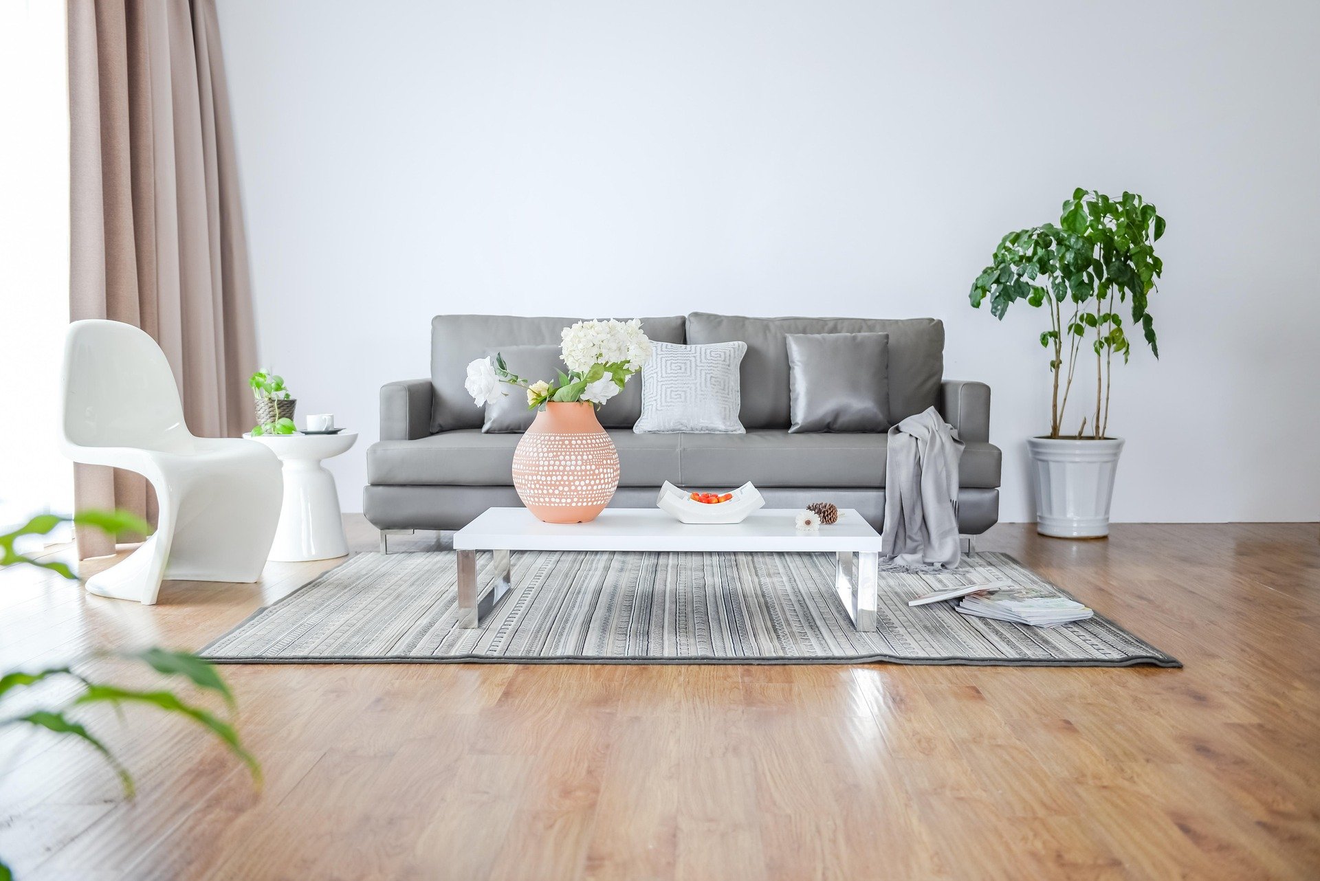 A modern living room with a gray sofa, white coffee table, white and gray pillows, a large potted plant on the right, beige curtains on the left, a patterned area rug, decorative flowers in a peach vase, and some magazines on the floor.