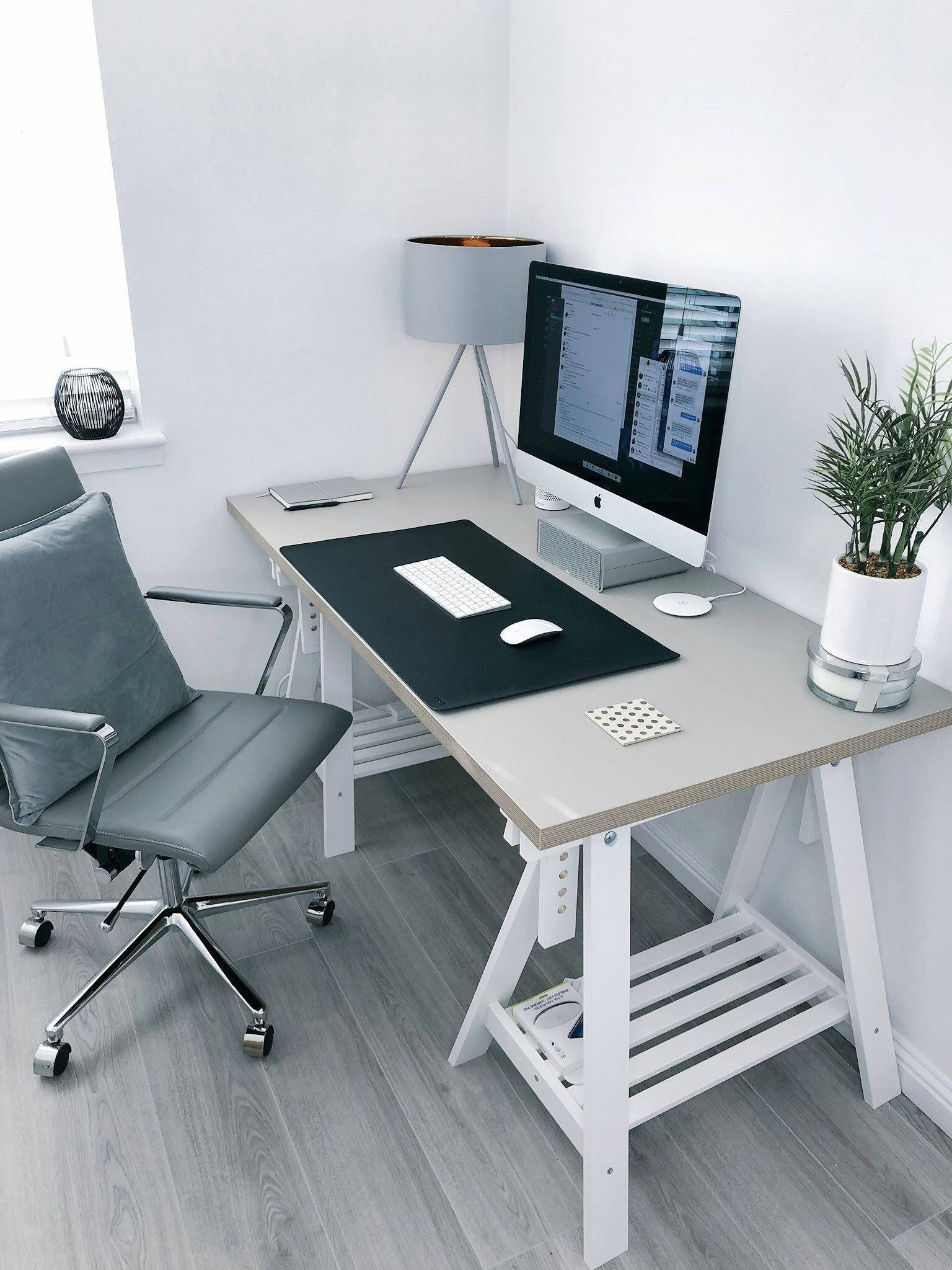 A modern workspace with a white desk, a computer monitor displaying messages, a wireless keyboard and mouse, a gray office chair, a table lamp, a small black decorative item, and a potted plant on the right side.