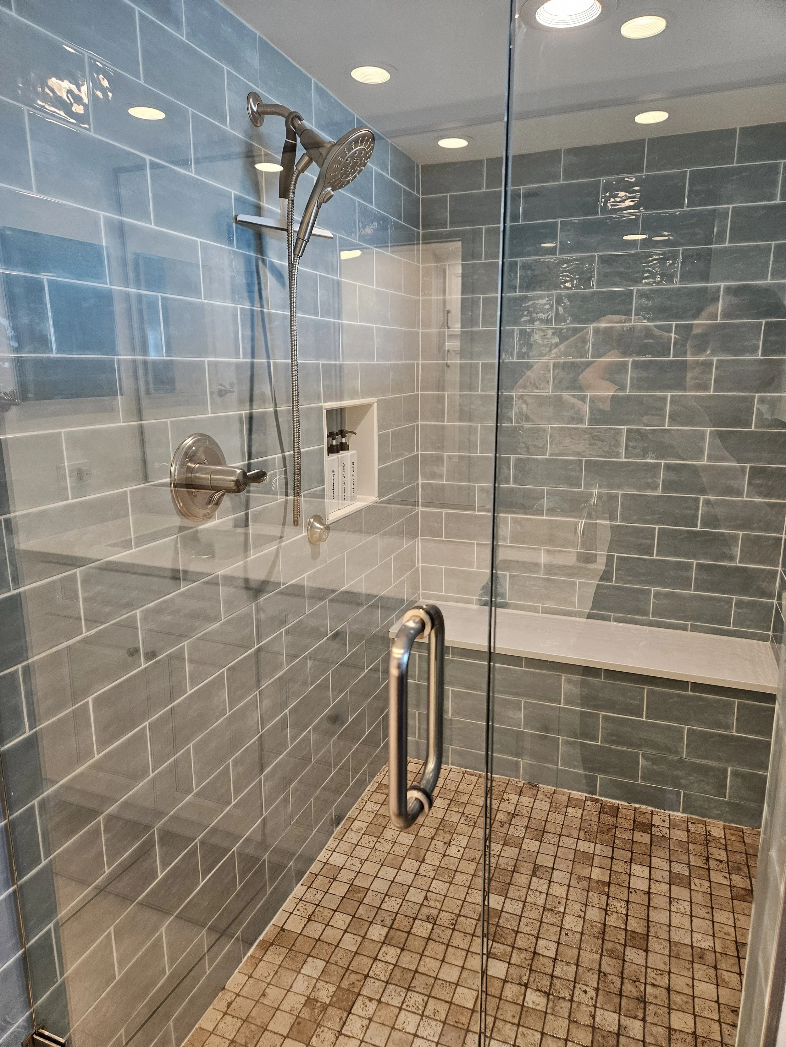 Inside a modern shower stall with blue-gray tiled walls, a handheld showerhead on an adjustable rail, and a recessed shelf with bottles. The floor has small square beige tiles, and the shower is enclosed with a glass door with a metal handle.