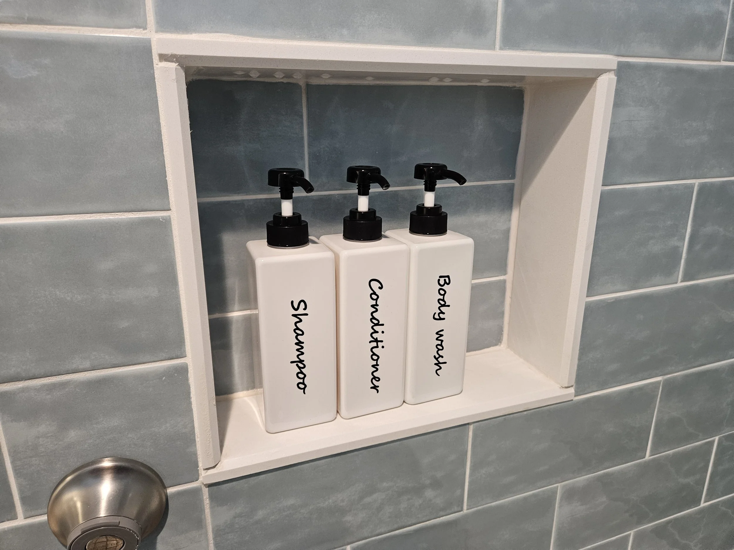 Airbnb restocked amenities. Bathroom shelf with three bottles labeled 'Shampoo', 'Conditioner', and 'Body Wash' next to a silver showerhead.