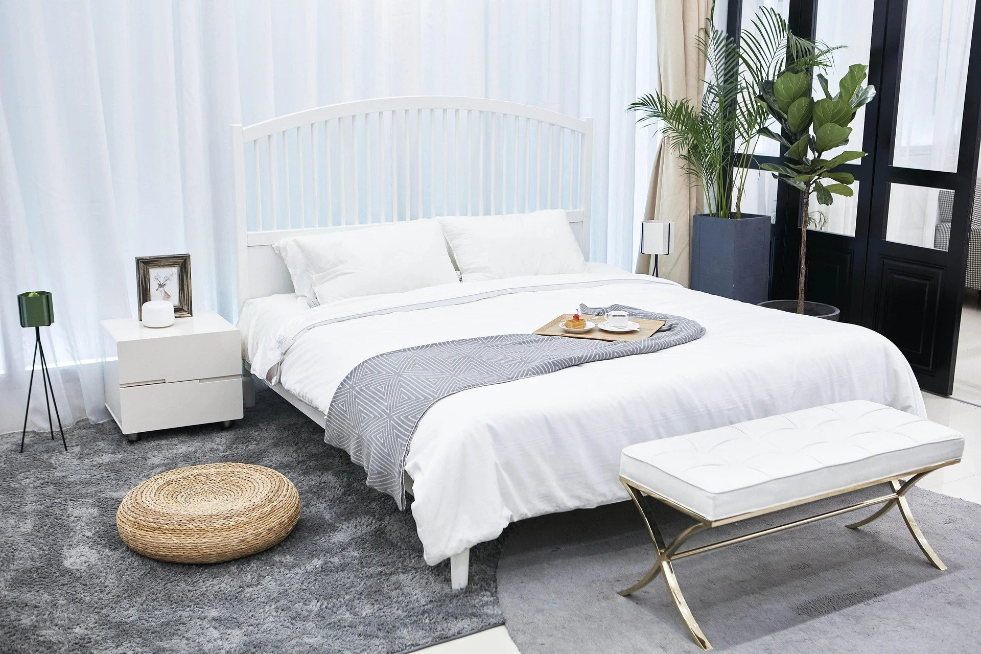 Modern bedroom with white bed, nightstand, tray with breakfast, gray rug, plant, bench, and black sliding door.