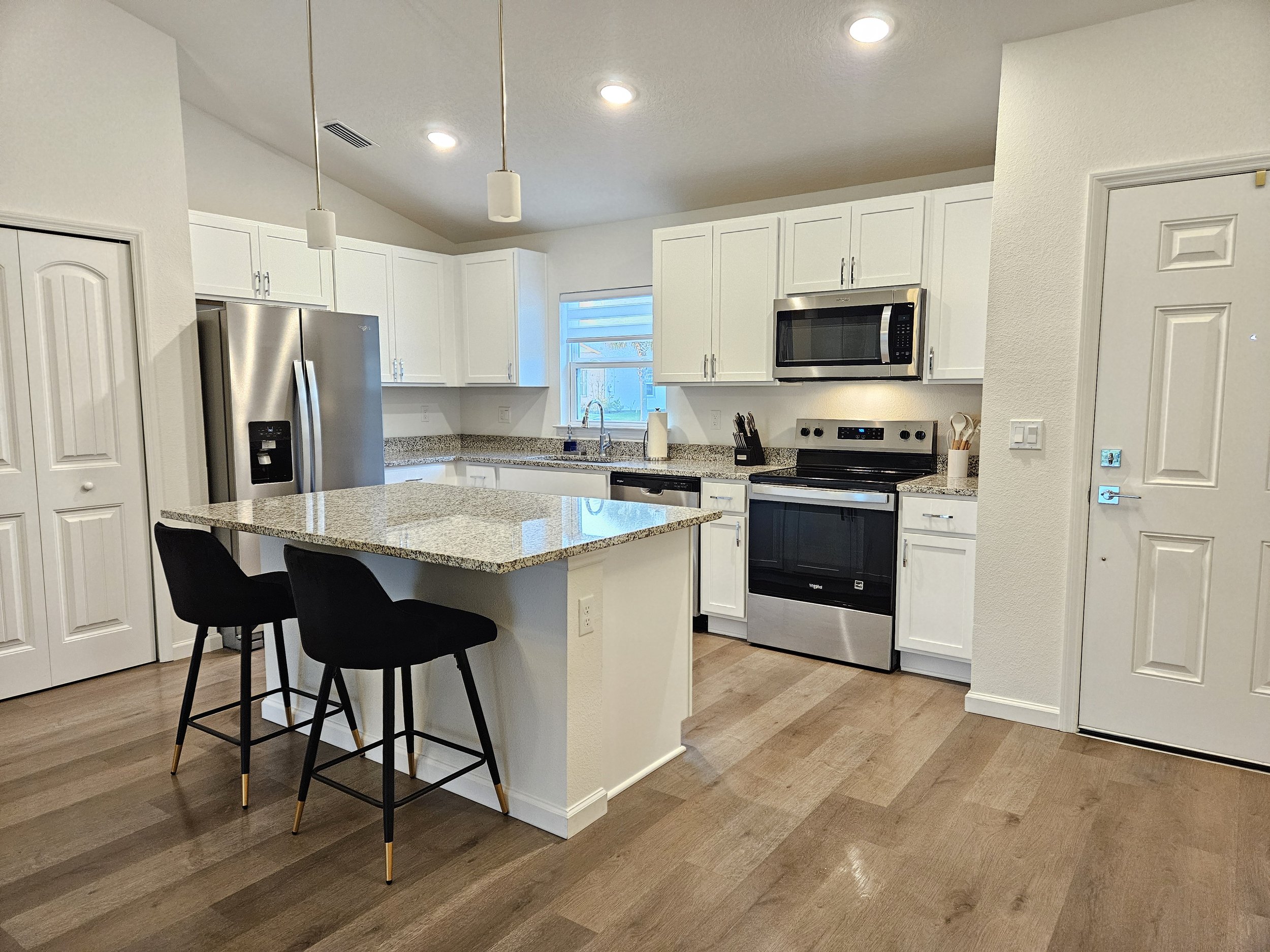 Modern kitchen with white cabinets, stainless steel appliances, a granite countertop island, and black bar stools. There is a window above the sink, and the floor is wooden.
