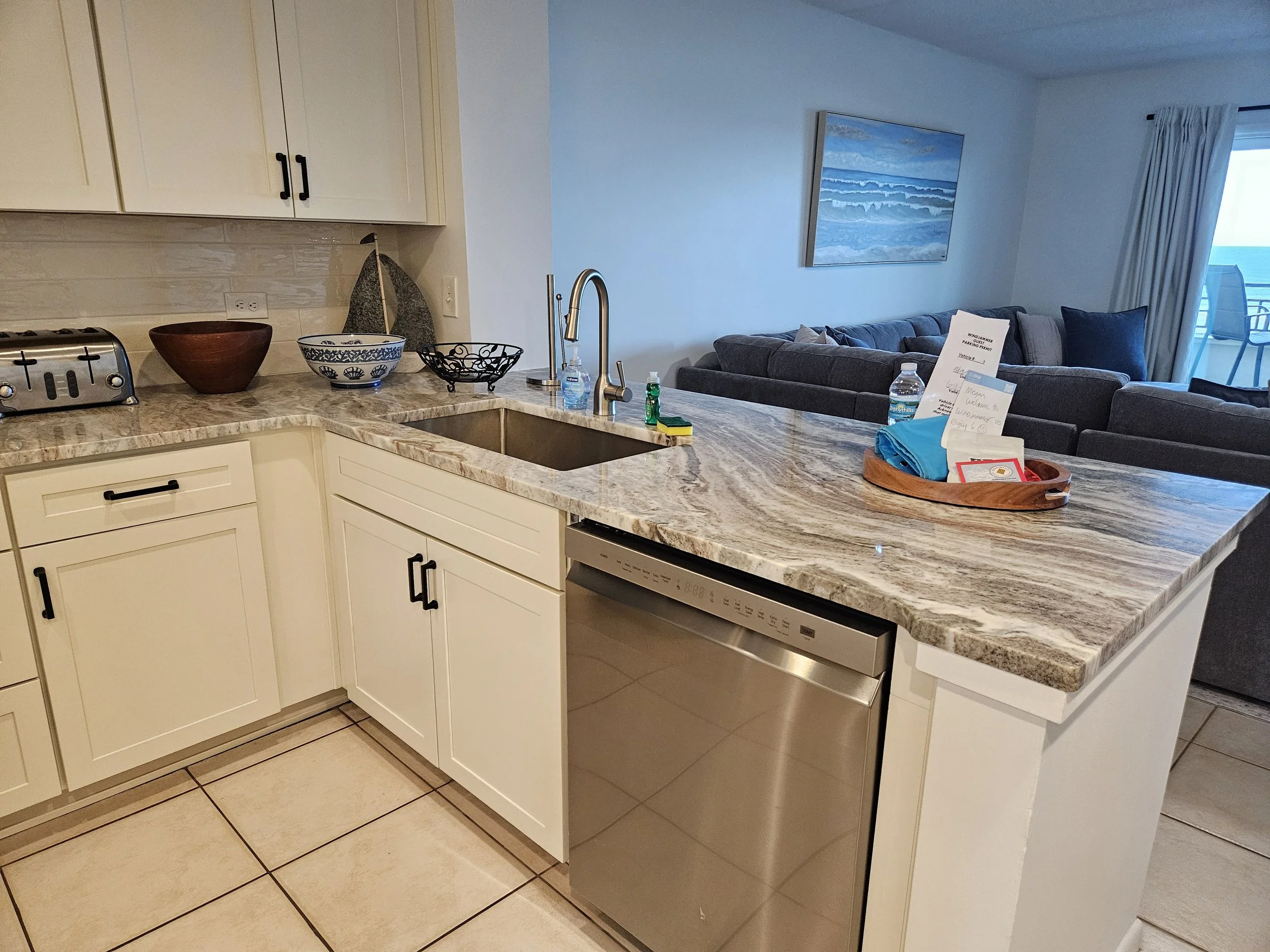 Kitchen area with white cabinets, a marble countertop, and a stainless steel dishwasher. Items on the counter include a wooden bowl, a decorative bowl, a black wire basket, soap and sanitizer dispensers, and a sponge. The background shows a living ro