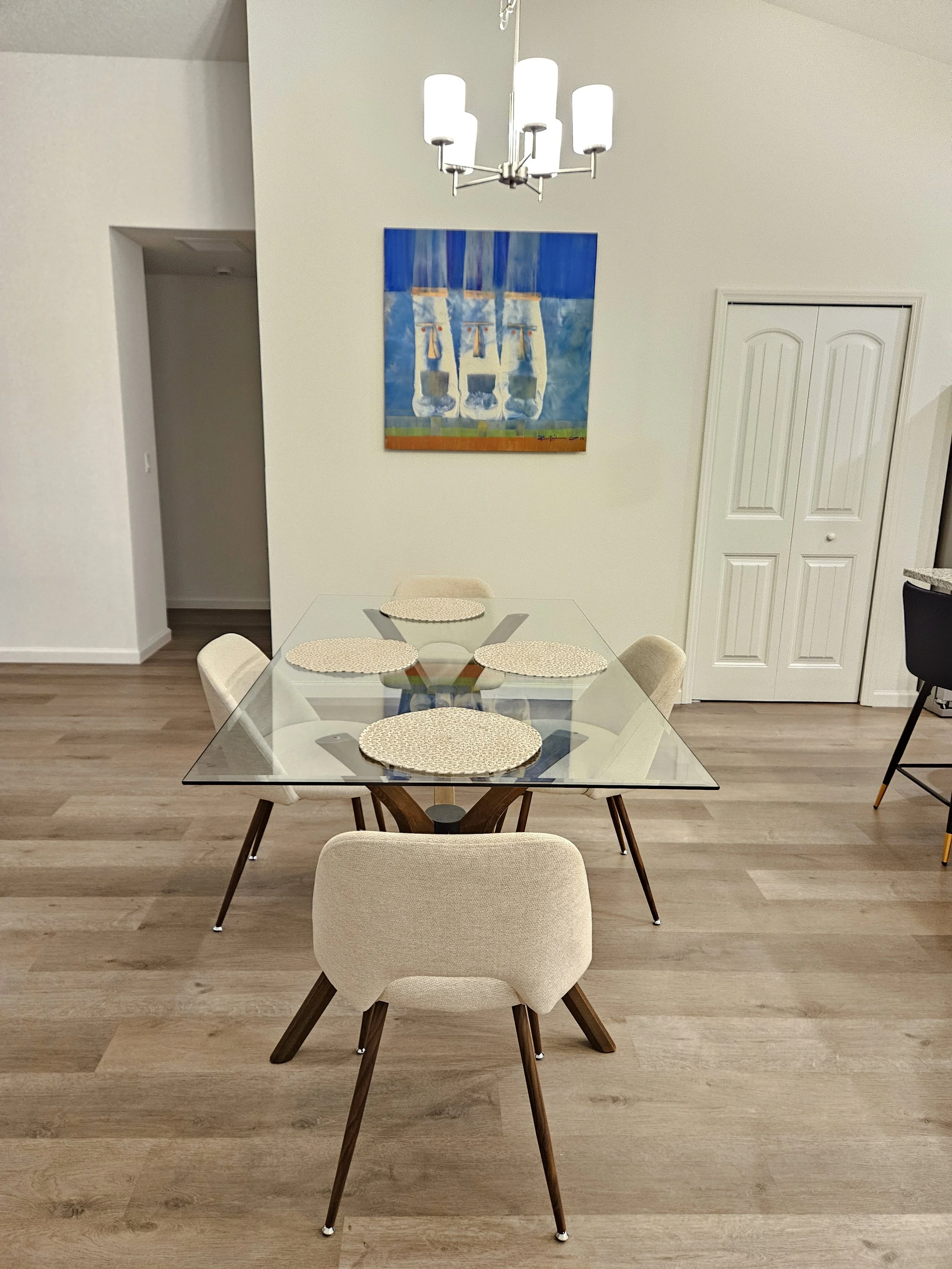 A dining room with a glass table and six beige chairs. There are four round placemats on the table. A chandelier hangs above, and a colorful abstract painting is on the wall behind the table.