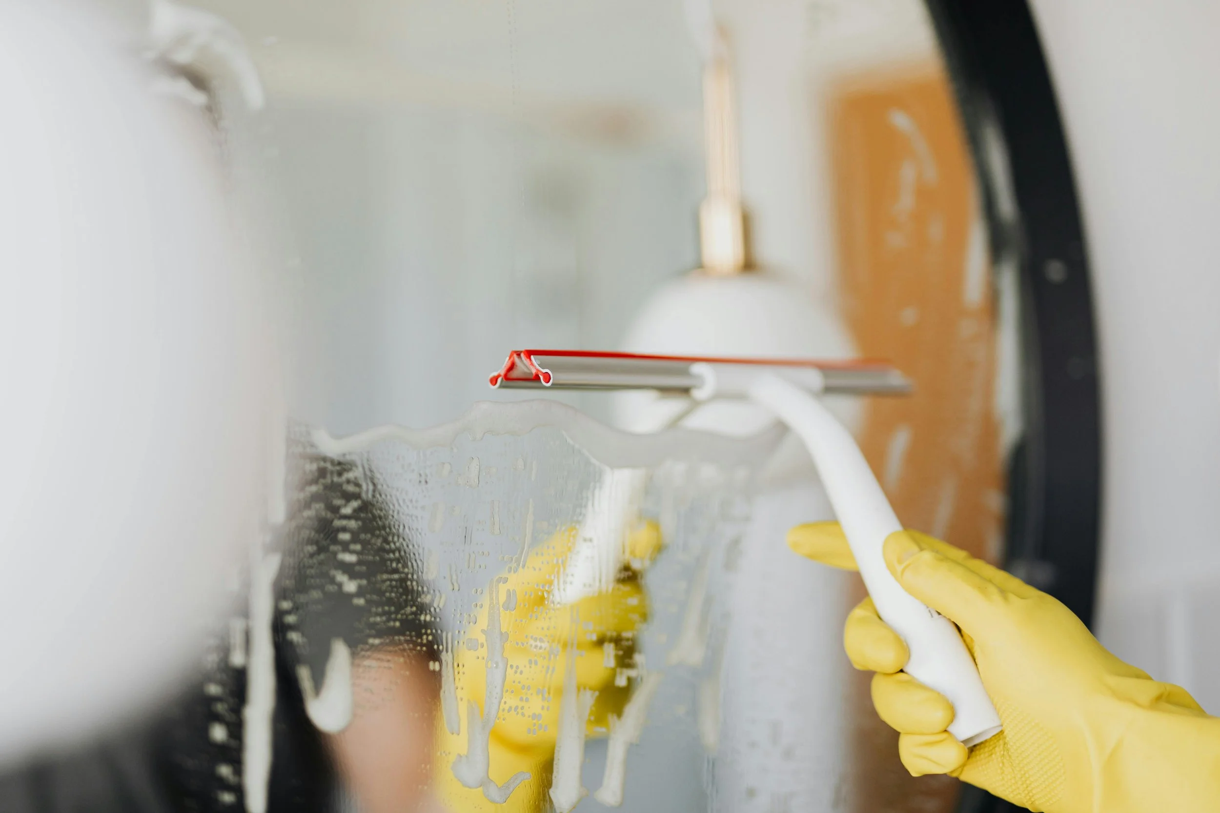 A person wearing yellow gloves cleaning a glass window with a squeegee, with soap suds on the glass.