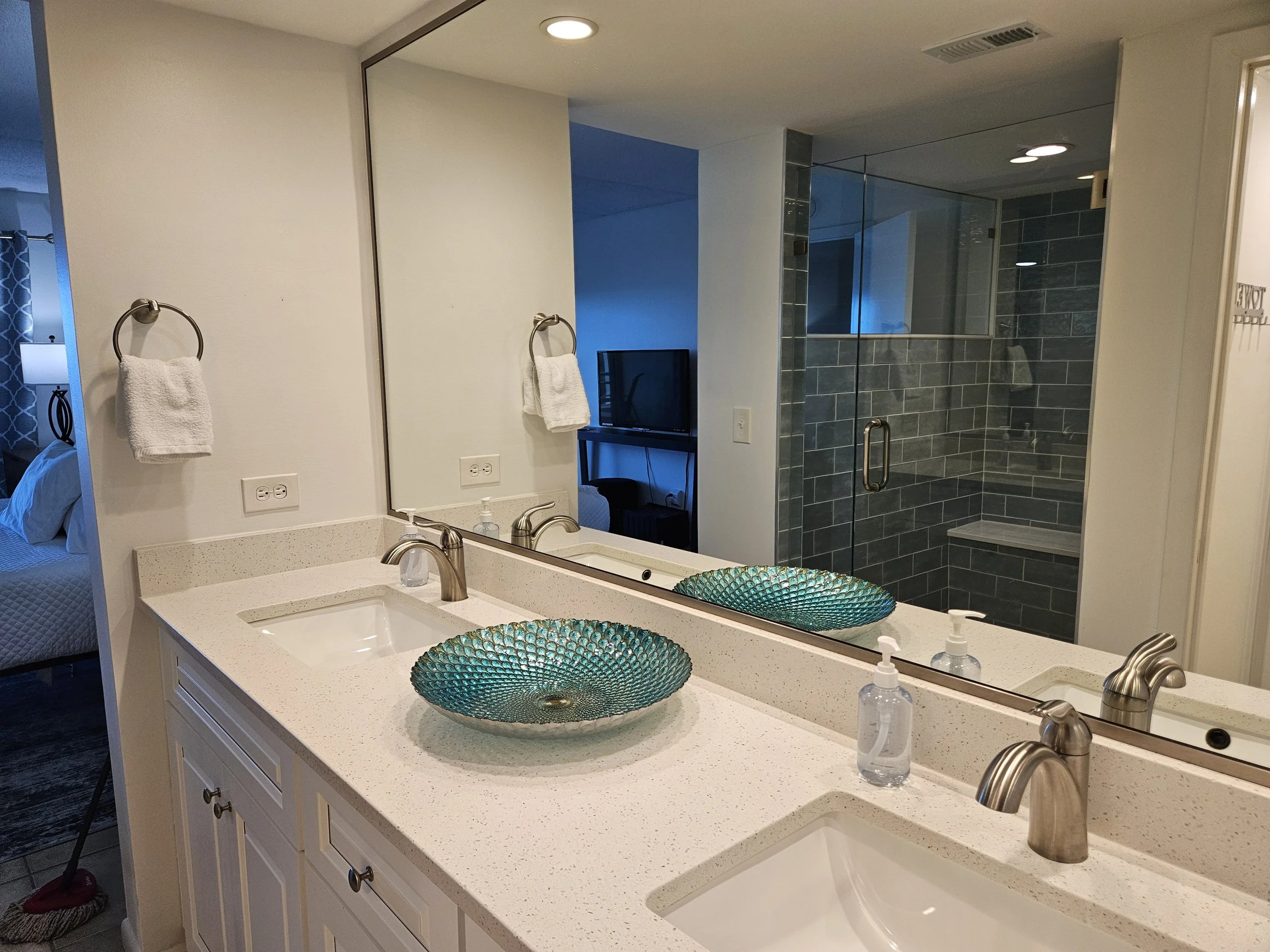 A bathroom vanity with two sinks, each decorated with a colorful, textured ceramic basin. The vanity has a large mirror above it, and a soap dispenser and a hand soap bottle are on the countertop. There are two hand towels hanging on the wall. A show