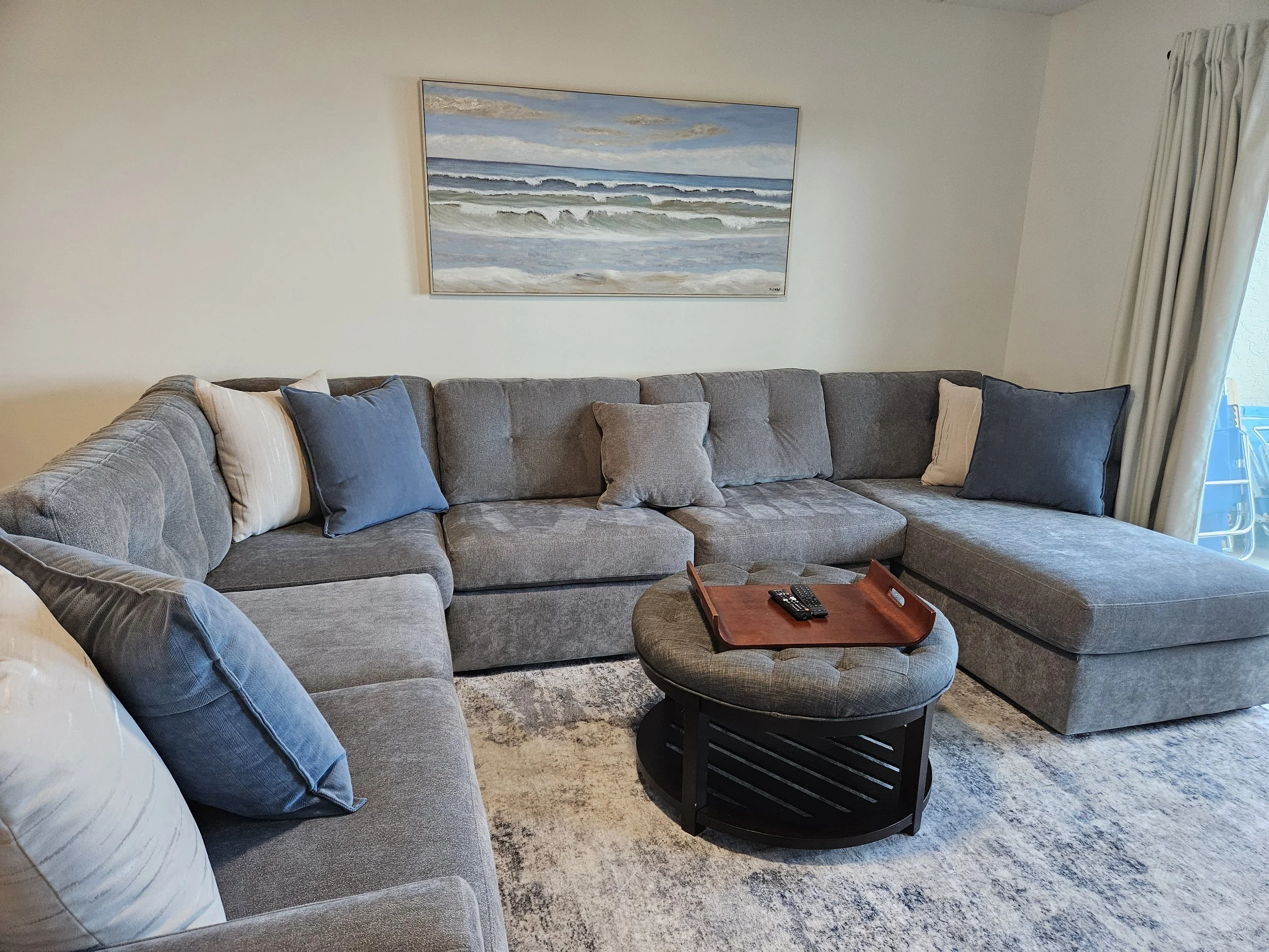 An airbnb staged Living room with gray sectional sofa, various pillows, a round ottoman with a tray and remote control, a rectangular tray, a wall art of ocean waves, a window with curtains.