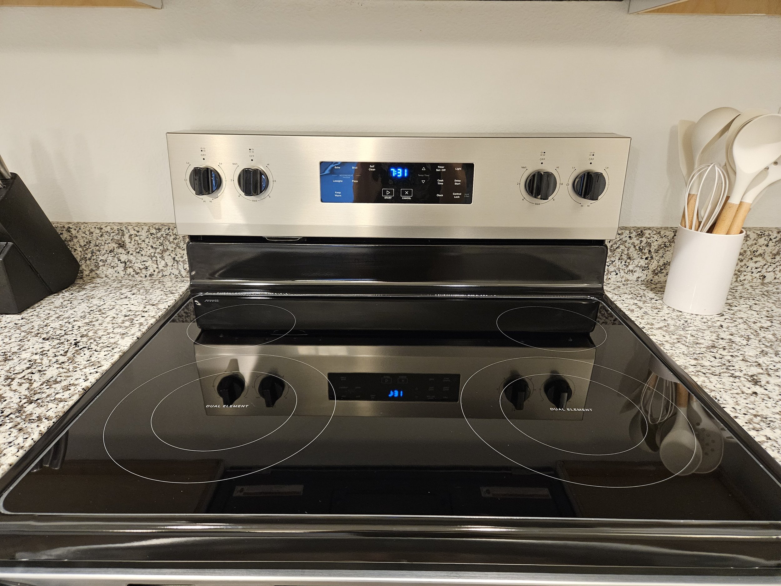 Kitchen stove with black ceramic cooktop and stainless steel control panel, white wall and granite countertop, utensil holder with white and wooden spoons.