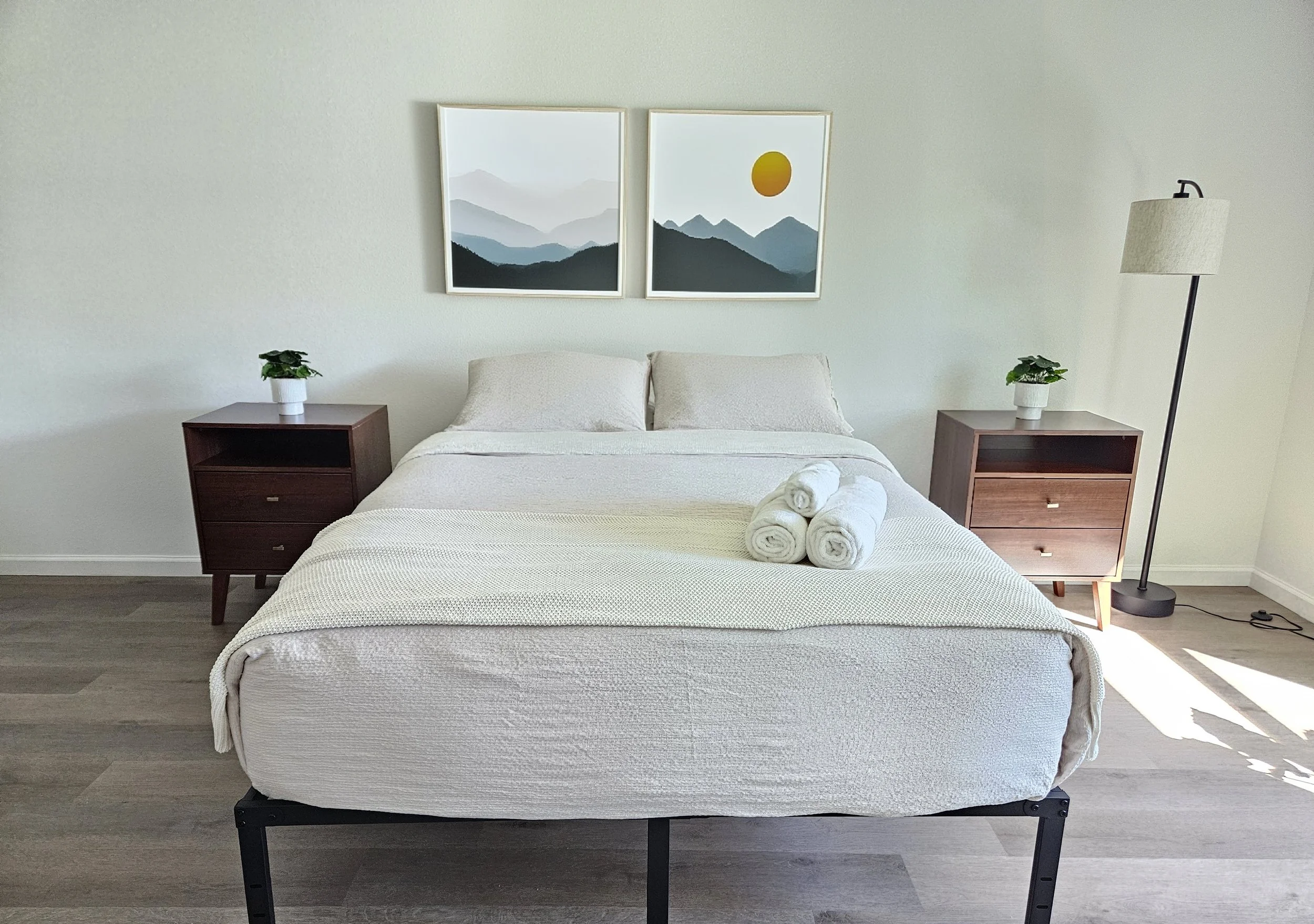 A neatly made bed with white bedding and three rolled towels on top, flanked by two dark brown nightstands with small potted plants, in a minimalist bedroom with a neutral-colored wall and artwork of mountain landscapes and a sun.