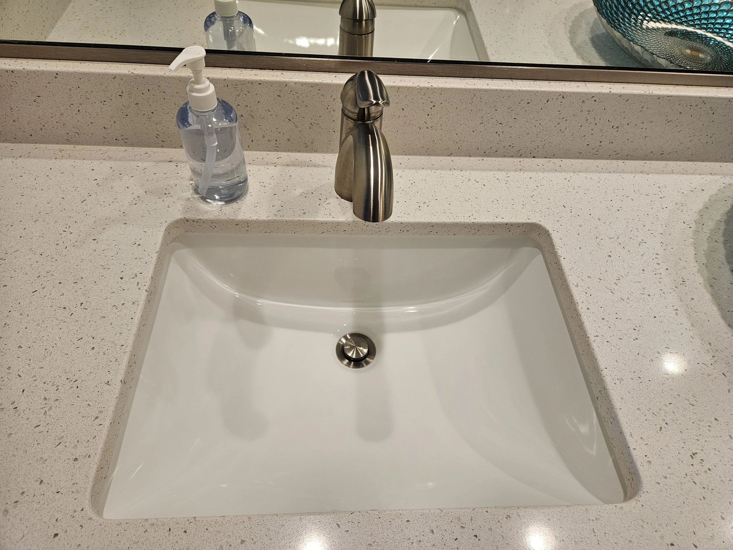 Bathroom sink with a chrome faucet, a bottle of hand sanitizer with a pump, and a large mirror reflecting part of the countertop and a blue decorative bowl.