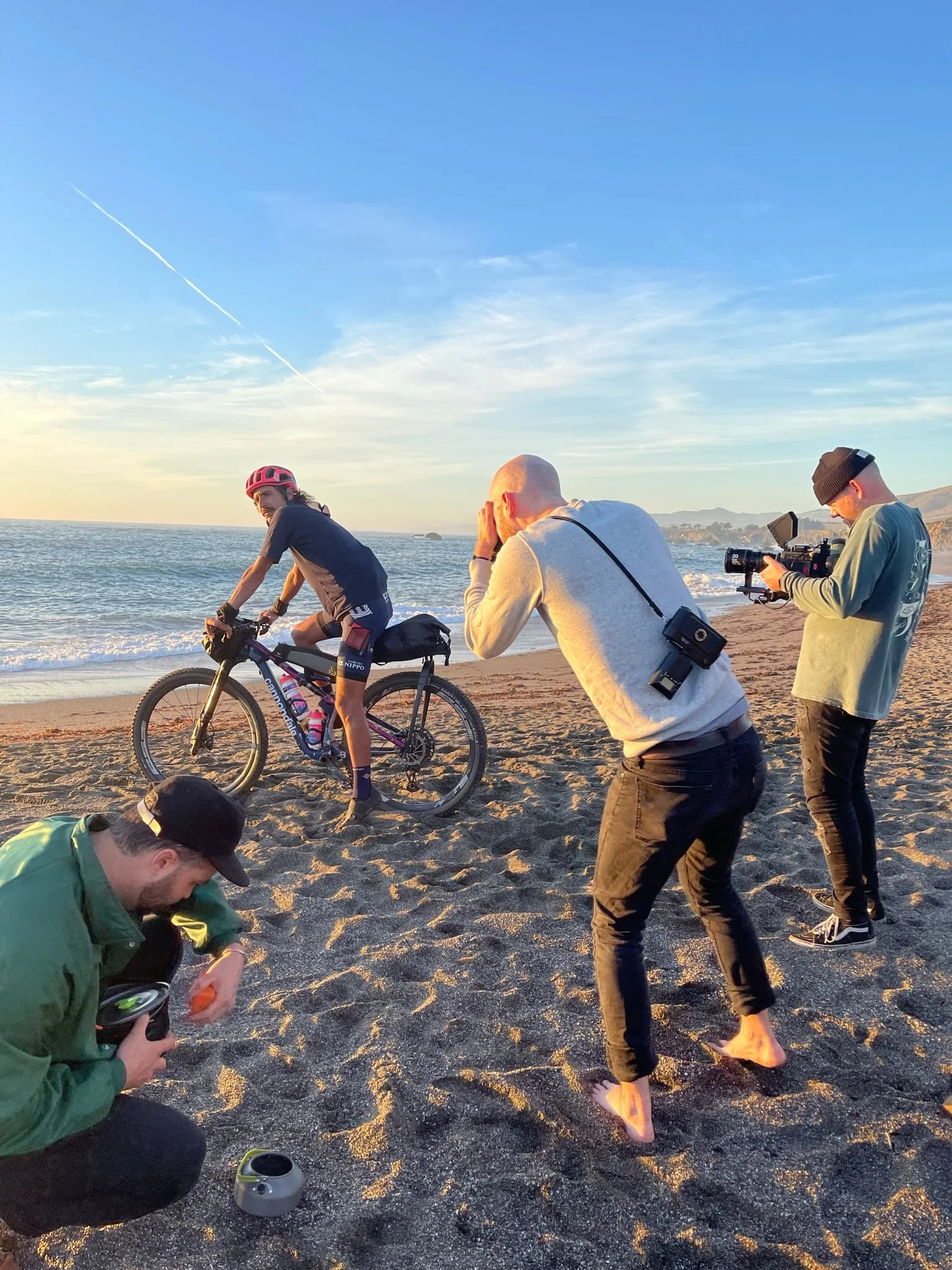 A group of people filming and taking photos on a beach with a cyclist riding near the shore at sunset.