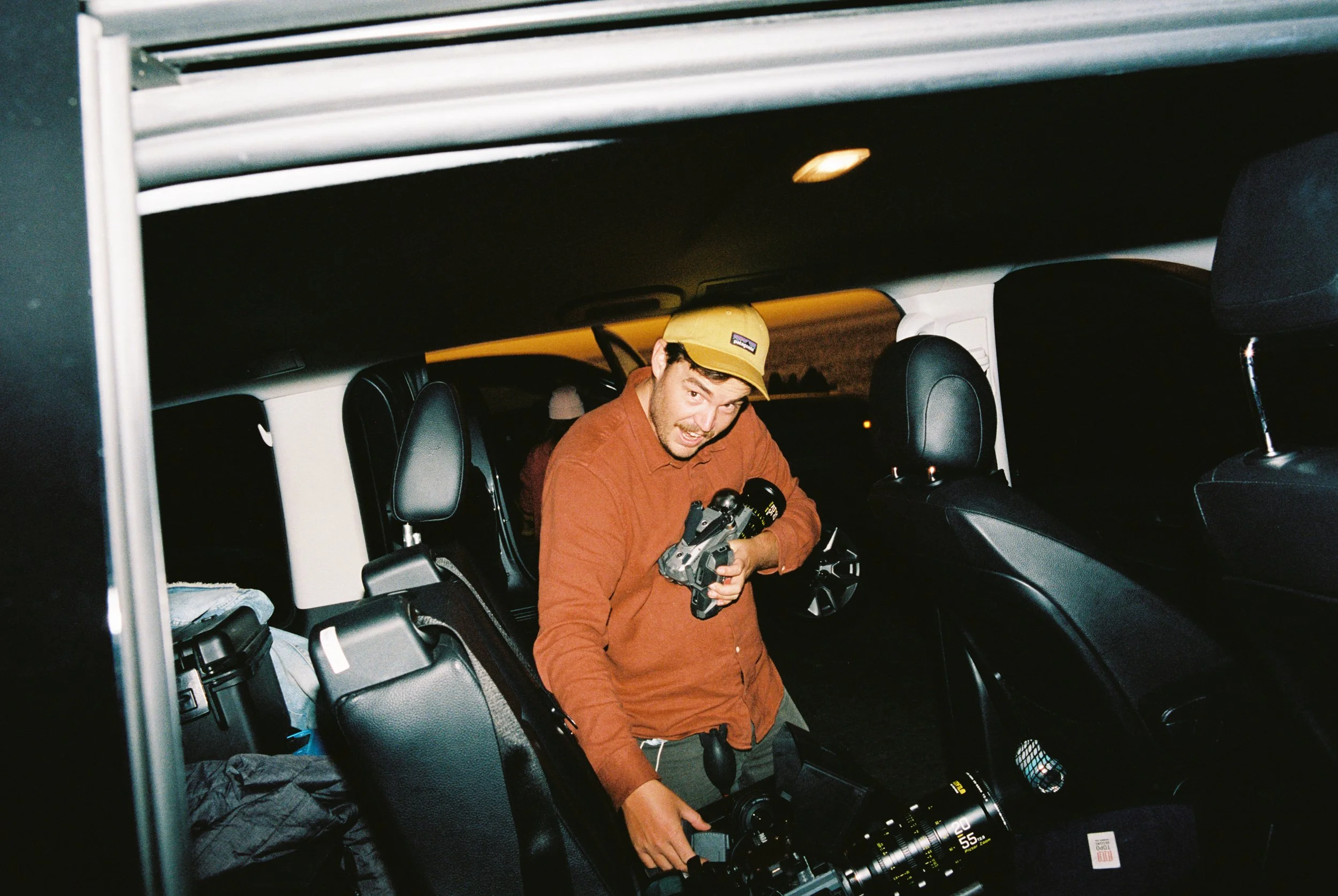 A man standing inside a vehicle at night, holding a camera and a video camera, with other equipment and gear around him.