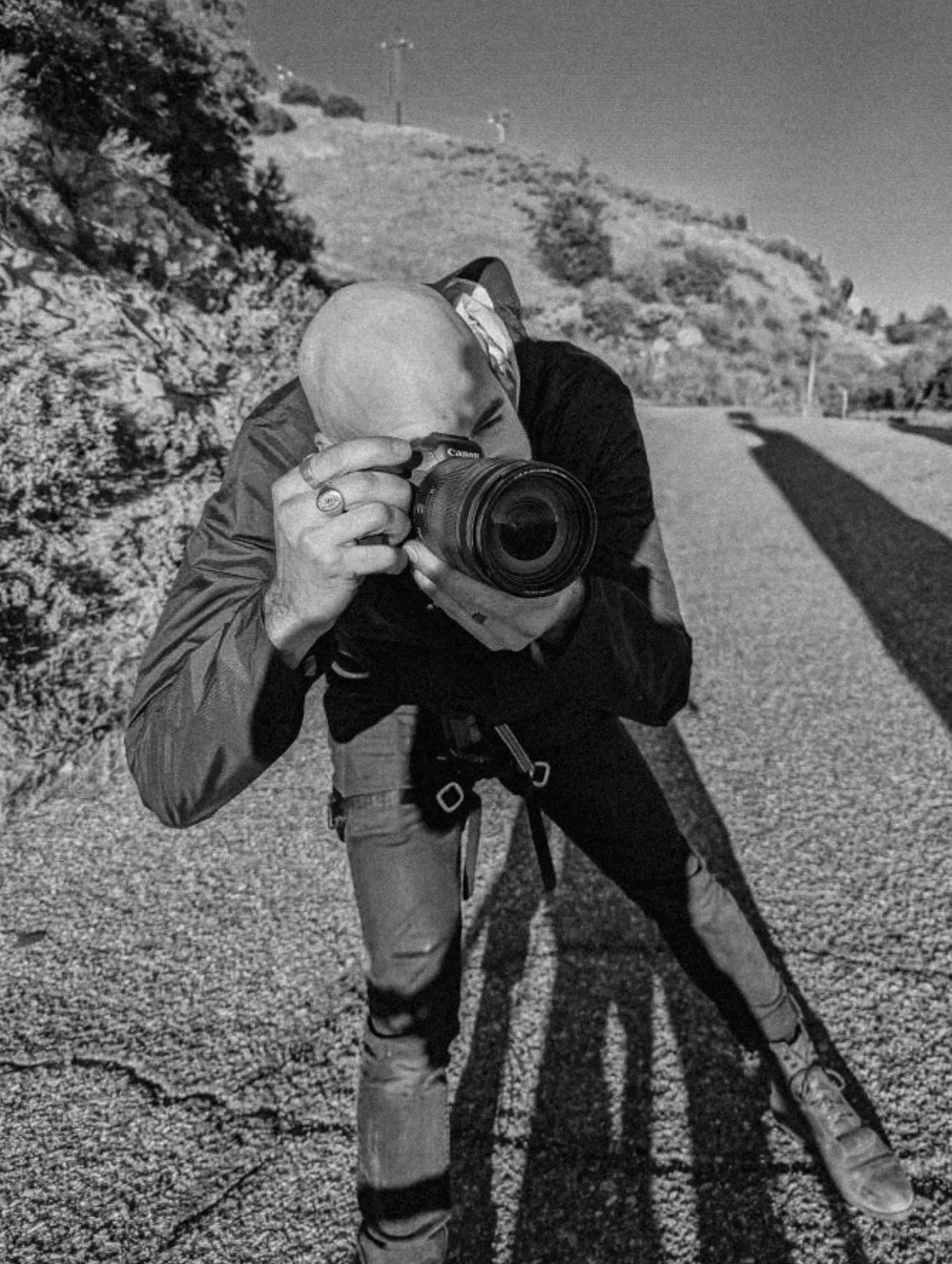 A man with a shaved head taking a photo with a digital camera on a road with hills and rocks in the background.