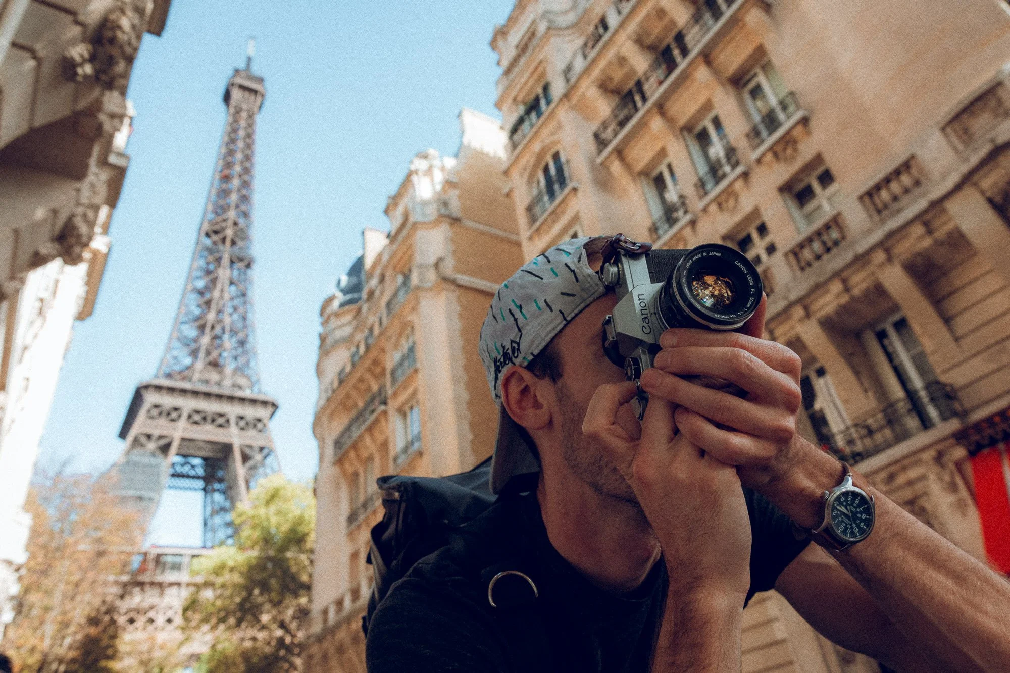 A man with a cap and watch is taking a photo with a camera on a city street, with the Eiffel Tower in the background.