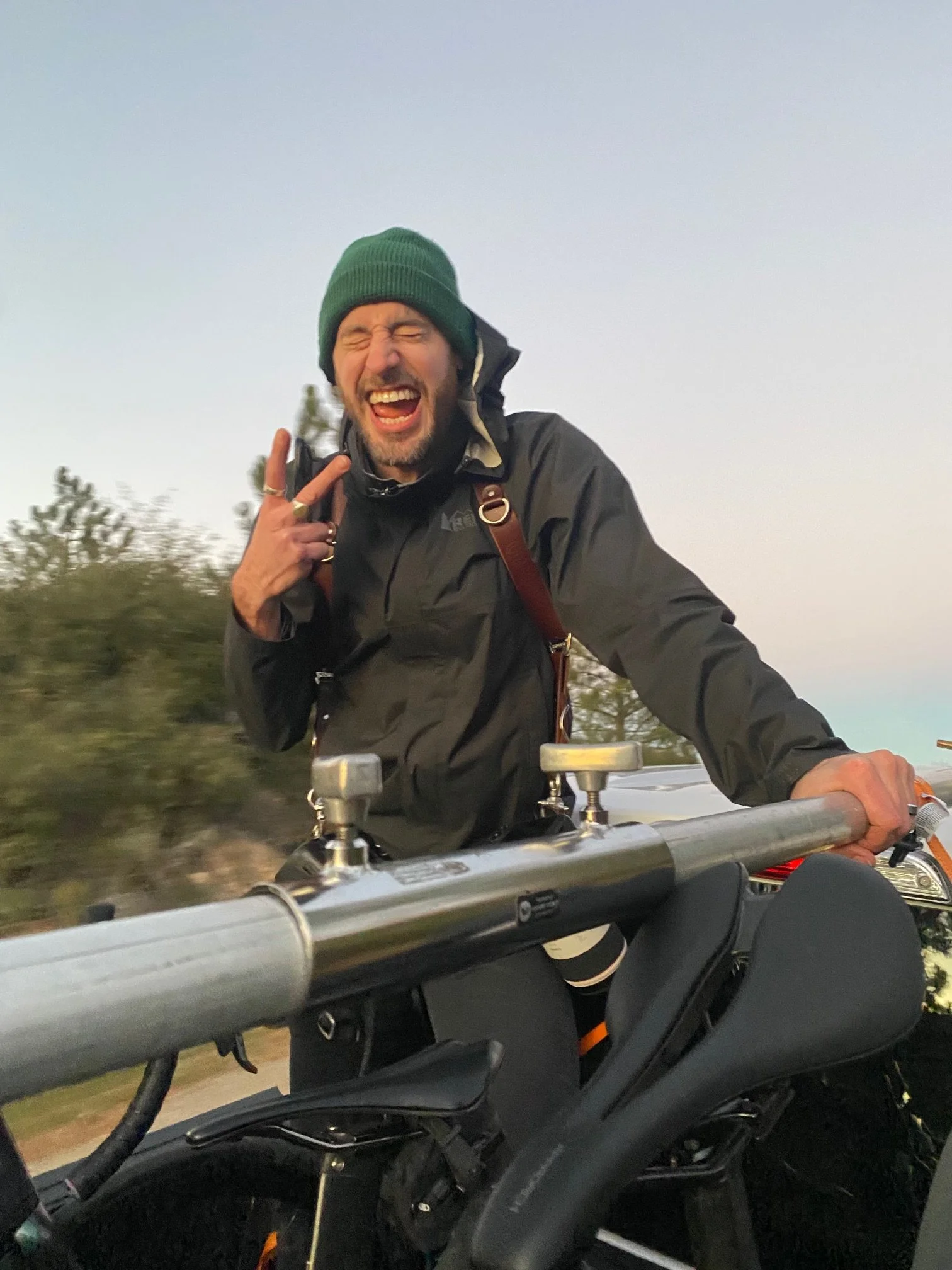 A man wearing a green beanie and a black jacket riding a motorcycle outdoors, making a peace sign and smiling with eyes closed.