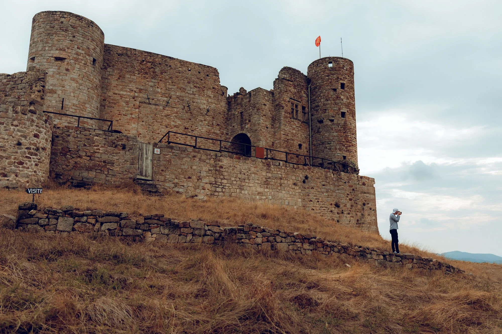 A person taking a photo of an old stone castle with a flag on top, on a grassy hill under a cloudy sky.