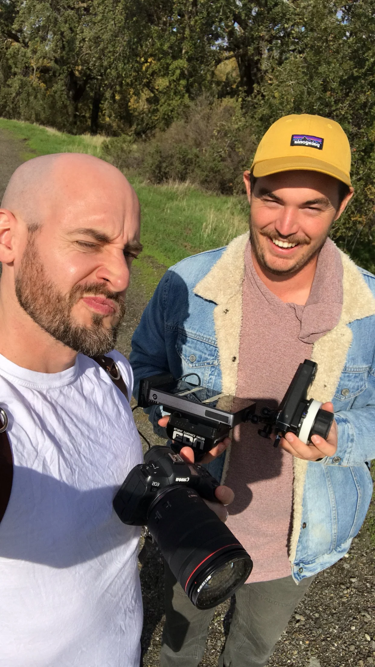 Two men holding camera equipment outdoors on a sunny day, one with a Canon camera and the other with a black device, standing on a dirt path with green grass and trees in the background.