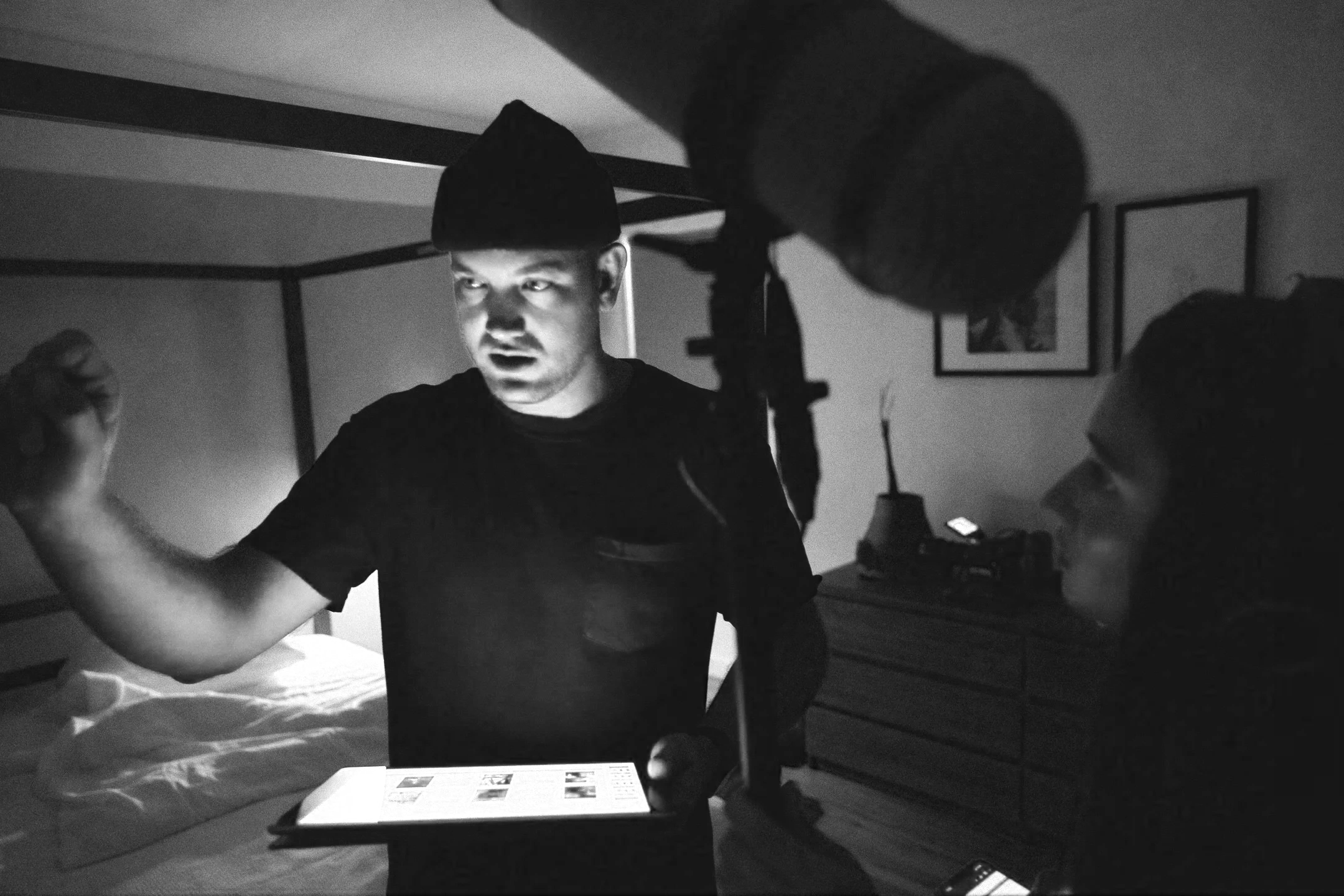 A man wearing a beanie hat is holding a tablet and appears to be directing an individual, possibly a woman, in a bedroom. The scene is dimly lit, and the photo is in black and white.