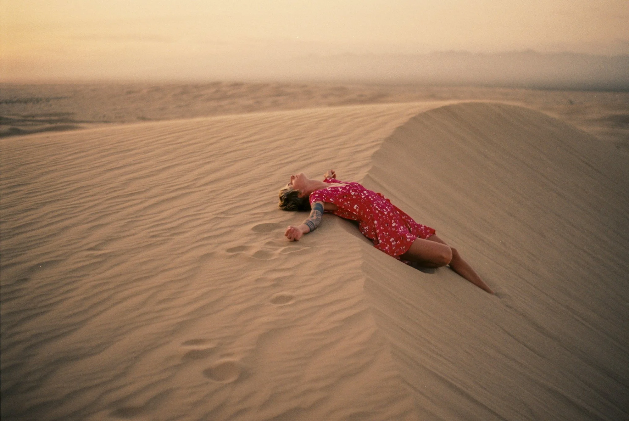 A woman lying on her back atop a sand dune in a desert, wearing a red dress with white floral pattern, with her arms and head tilted back, surrounded by sand with footprints nearby.