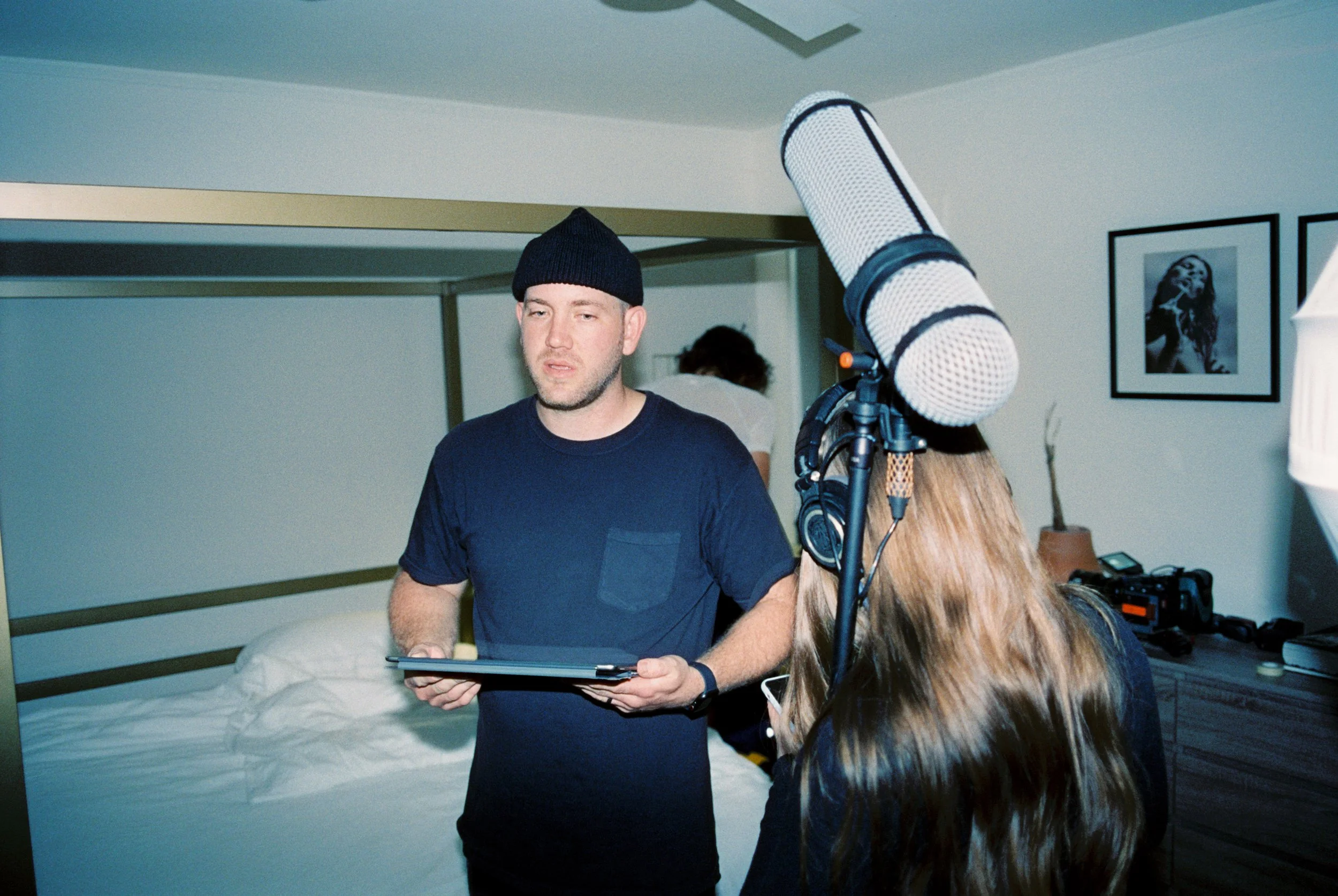 Filmmaker and crew filming in a bedroom, with a man holding a tablet and a woman with a microphone and headphones, and photographs on the wall.