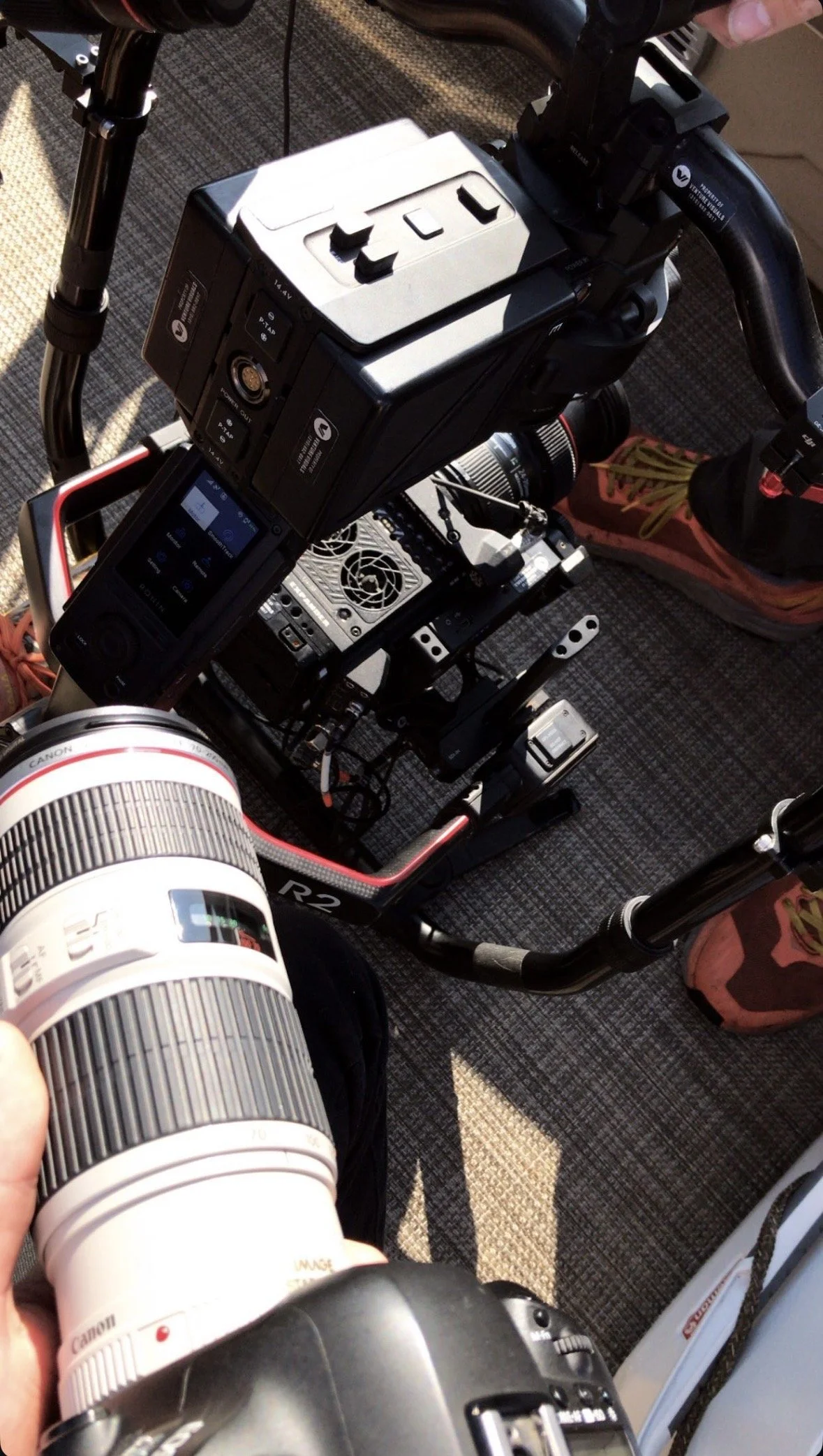 A video camera setup on a tripod, including a large white telephoto lens, a smaller black eyepiece, and various electronic equipment. The scene appears to be on a carpeted floor with a person's foot wearing a sneaker visible.