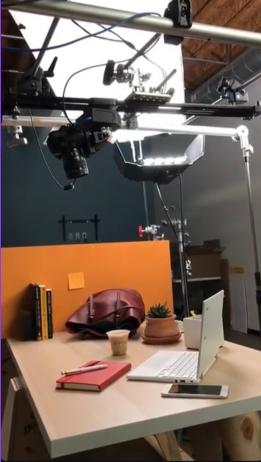 Camera and lighting equipment setup above a desk with books, a laptop, a phone, a purse, a plant, a cup, and notebooks.
