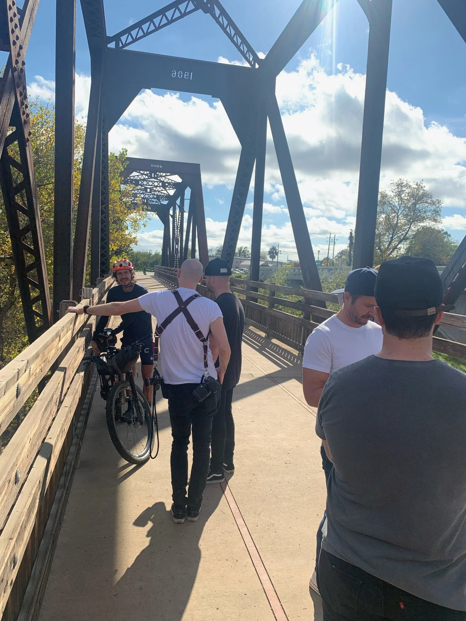 Group of people standing on a bridge under a blue sky with clouds. One person has a bicycle, others are talking and looking around.