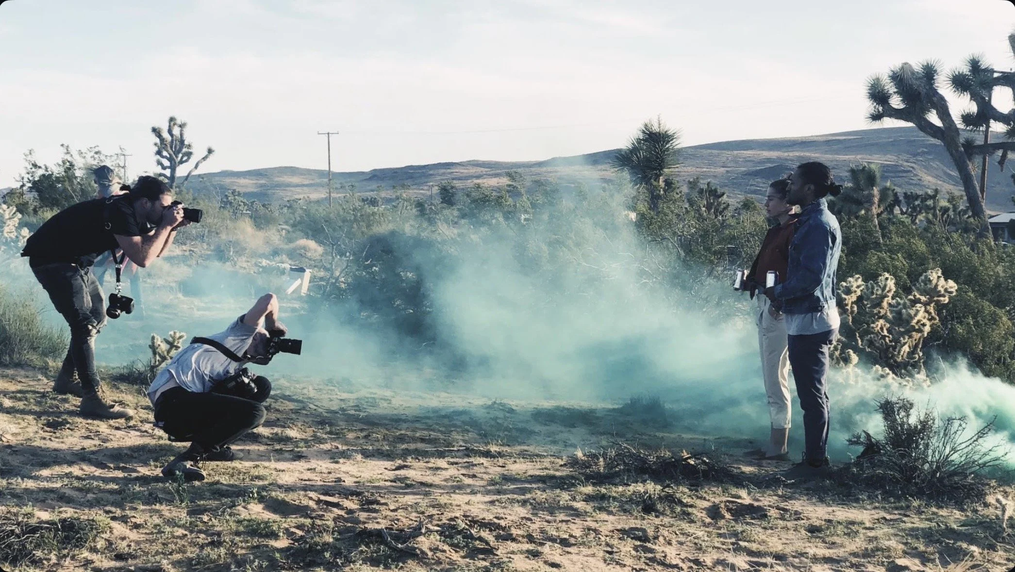 Two women are standing in a desert landscape with desert plants and a smoky haze around them. Two male photographers with cameras are capturing the scene.