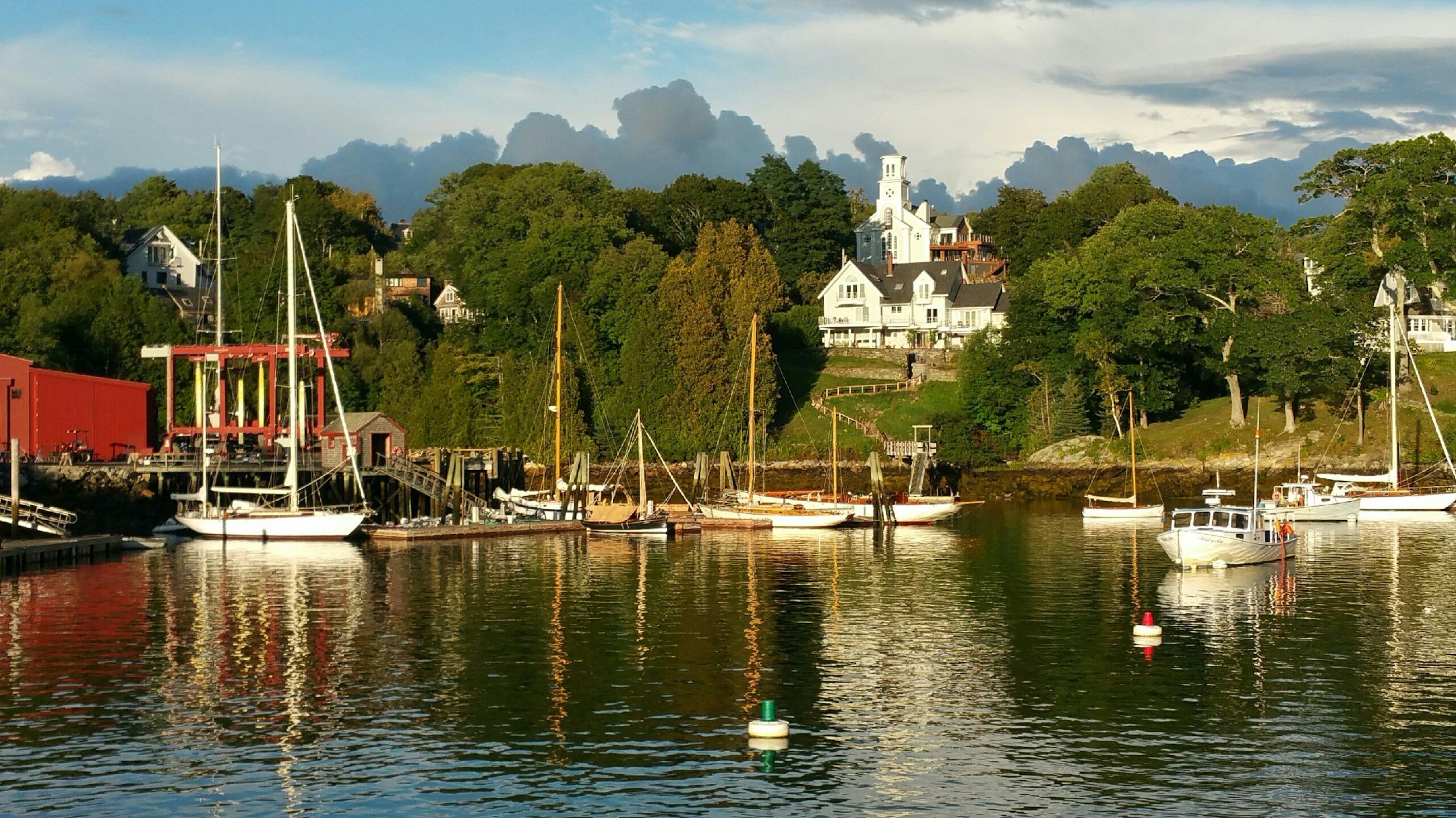 Boats docked at a marina with lush green trees and houses on a hill in the background under a partly cloudy sky.