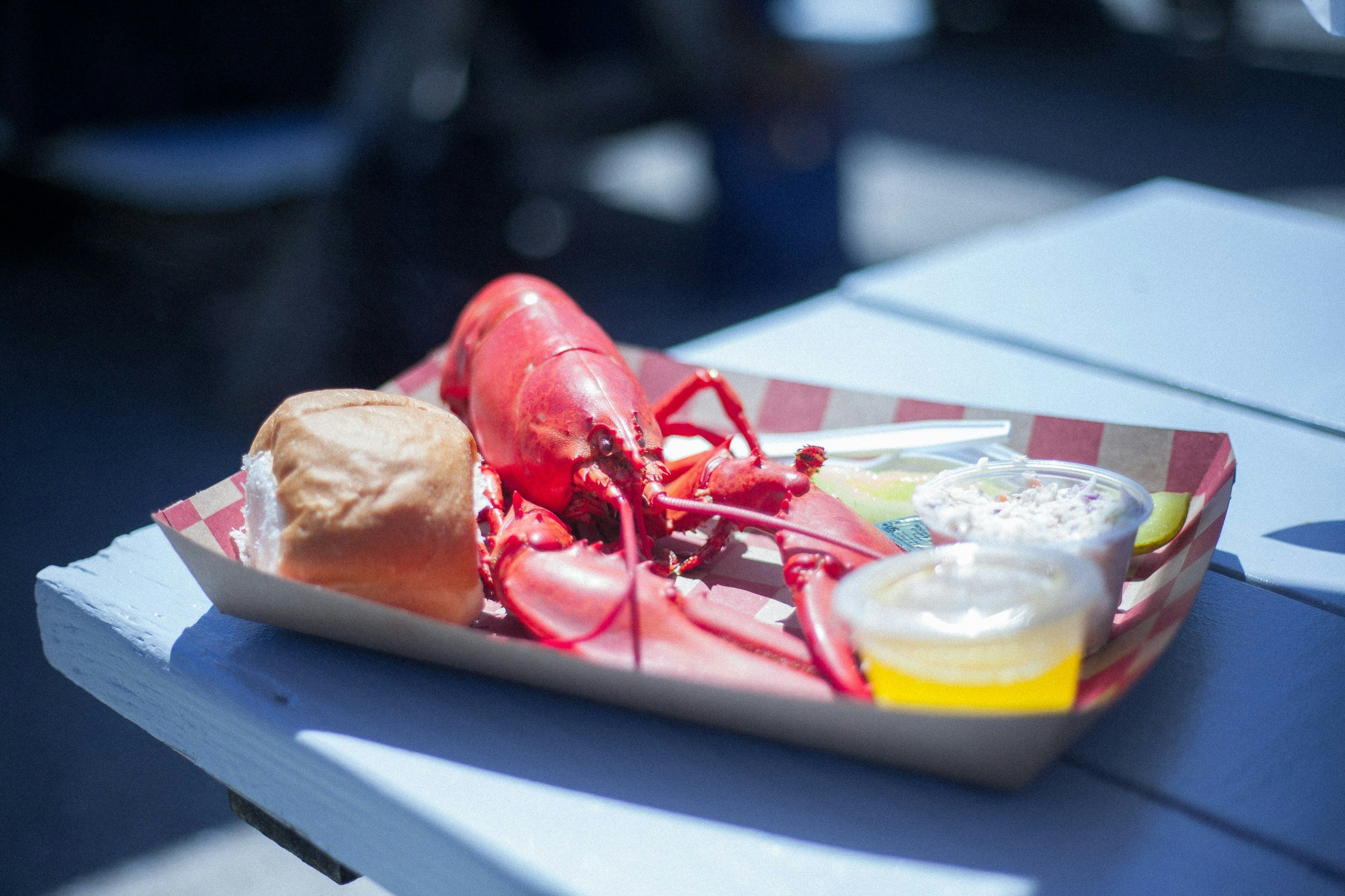 A food tray with a cooked lobster, a bread roll, and several small containers of sides and dipping sauces on a white wooden table outdoors.