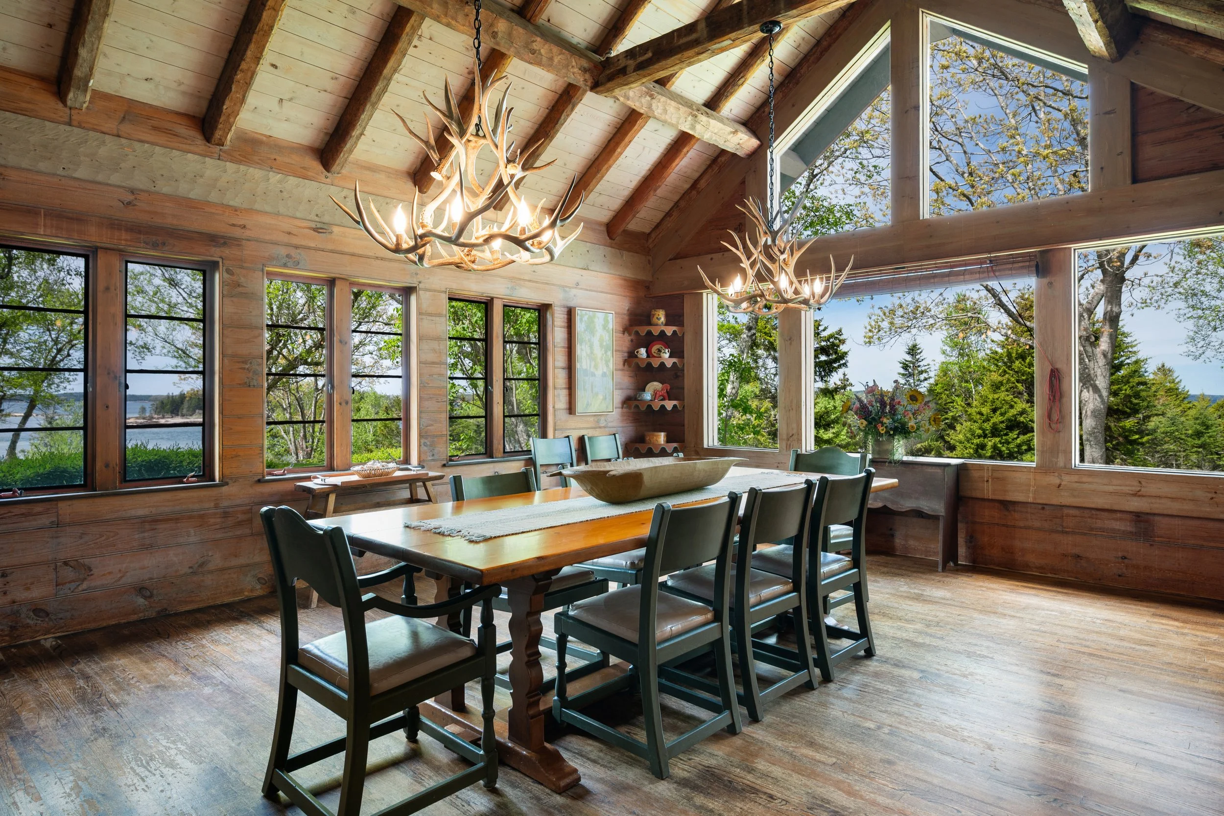 A rustic dining room with wooden walls and ceiling, large windows showing trees and a lake, a wooden dining table with chairs, and antler chandeliers hanging from the ceiling.