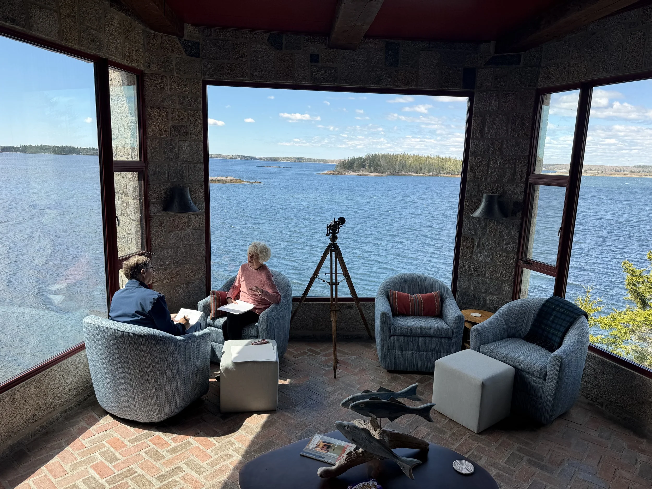 A cozy indoor space with large windows showing a scenic view of water, small islands, and a cloudy sky. Inside, there are several upholstered chairs and a small table with magazines, and a telescope is positioned near the window. Two people are seated and engaged in conversation.