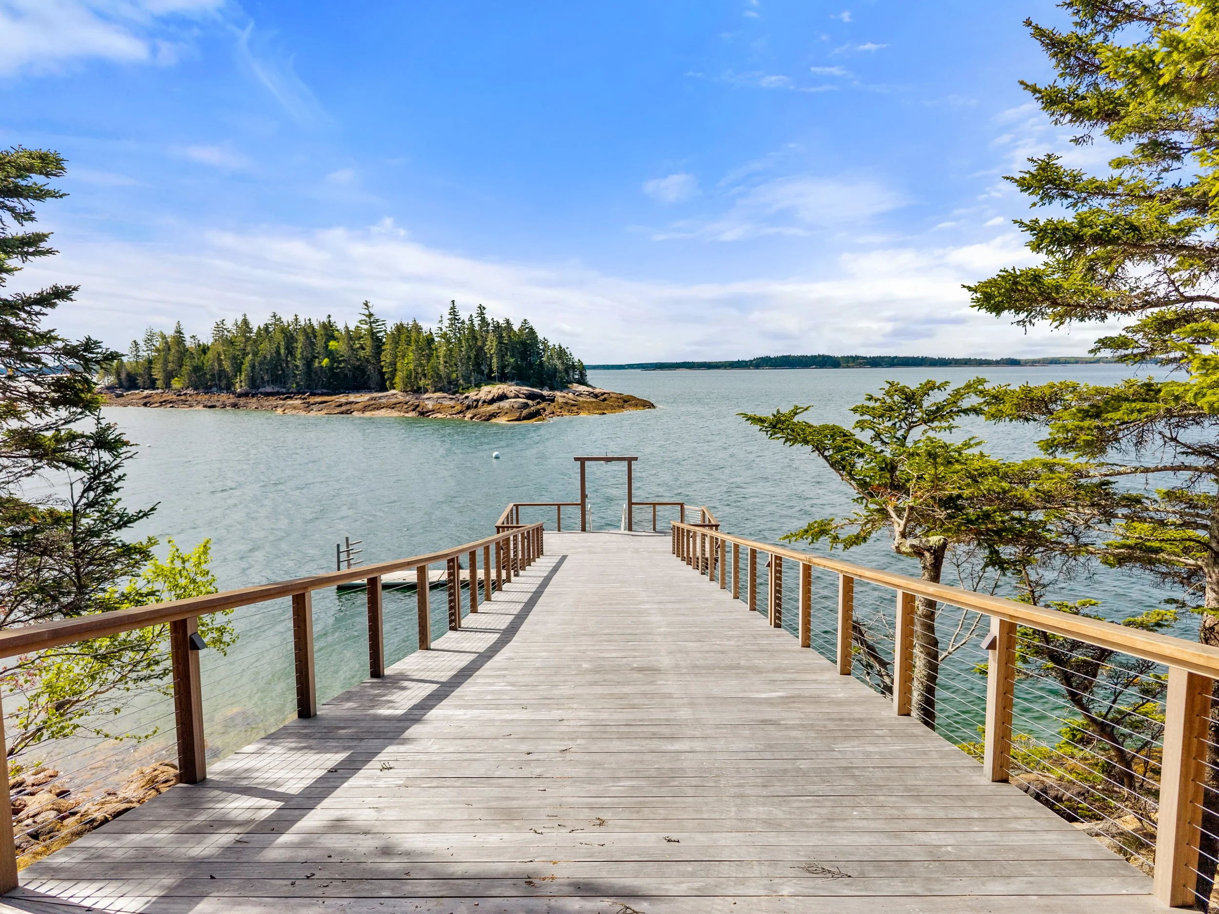 A wooden deck overlooks a body of water with a forested island and distant shoreline under a partly cloudy sky.