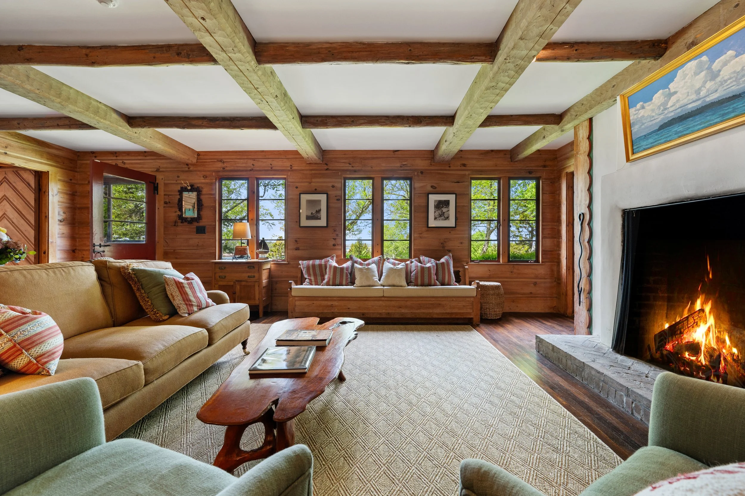 Living room with wooden walls, three large windows showing trees outside, a sofa with striped pillows, a coffee table with books, a fireplace with a fire, and framed artwork on the walls.