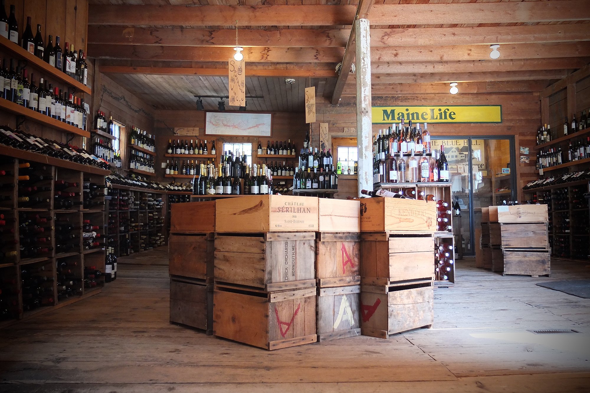 Blue Hill Wine Shop- Interior of a rustic wooden wine shop with shelves filled with wine bottles, a central wooden display with additional bottles, and a yellow sign reading 'MaineLife' on the back wall.