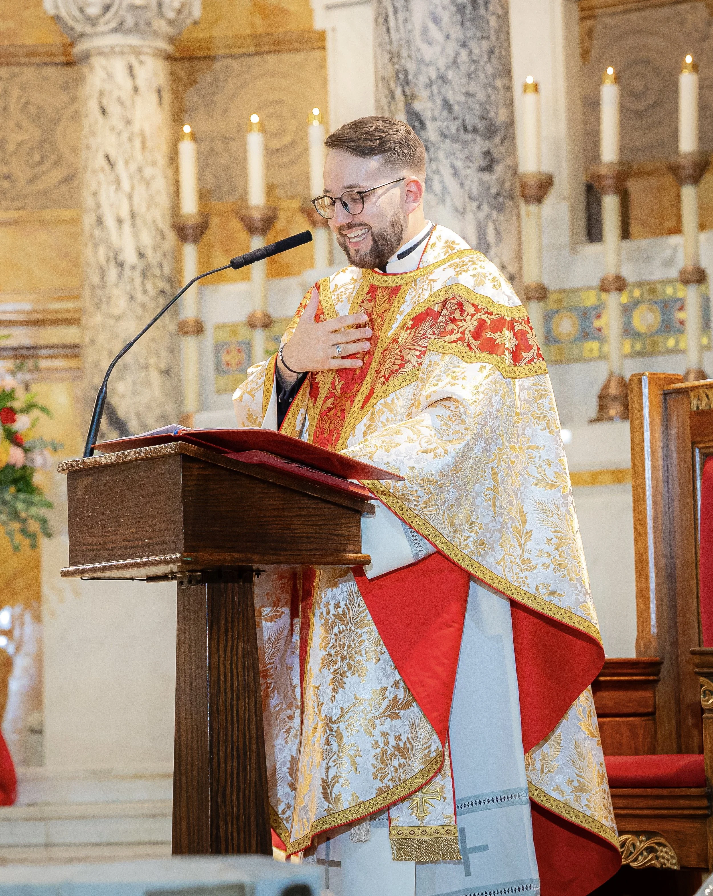 A priest dressed in ornate gold and red liturgical vestments stands at a wooden lectern inside a church, smiling and touching his chest with his right hand. The background shows church candles and marble columns.