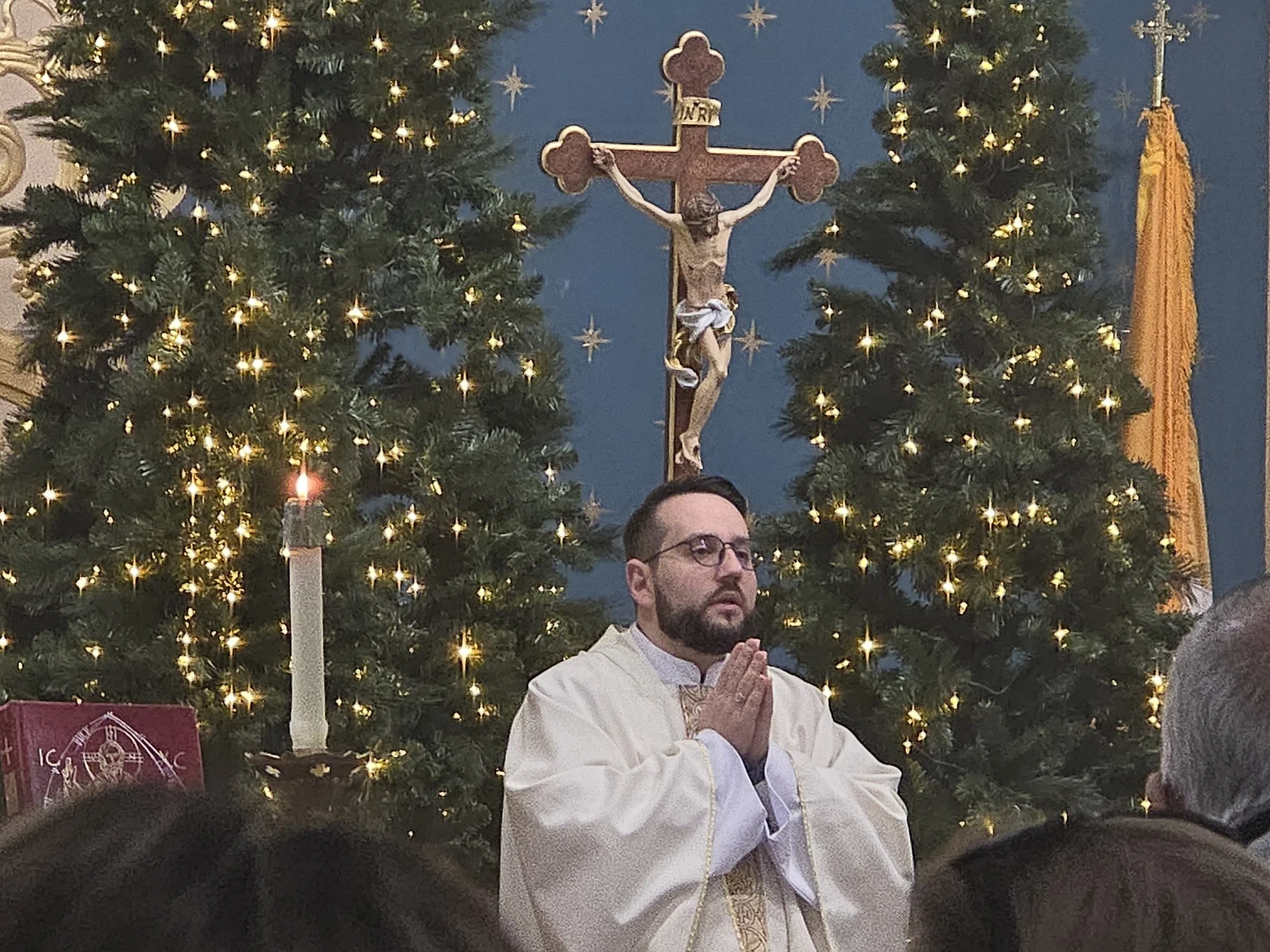 A man with glasses in a cream robe, praying with hands clasped, in front of Christmas trees with lights and a crucifix in the background.