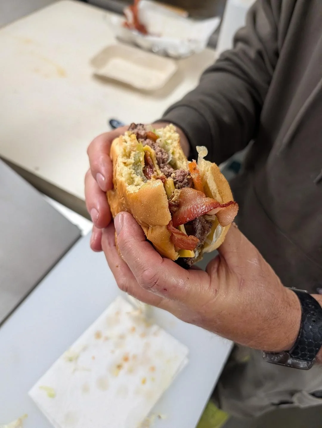 Person holding a partially eaten burger with visible bacon, beef patty, pickles, and other ingredients, in a kitchen or food preparation area.