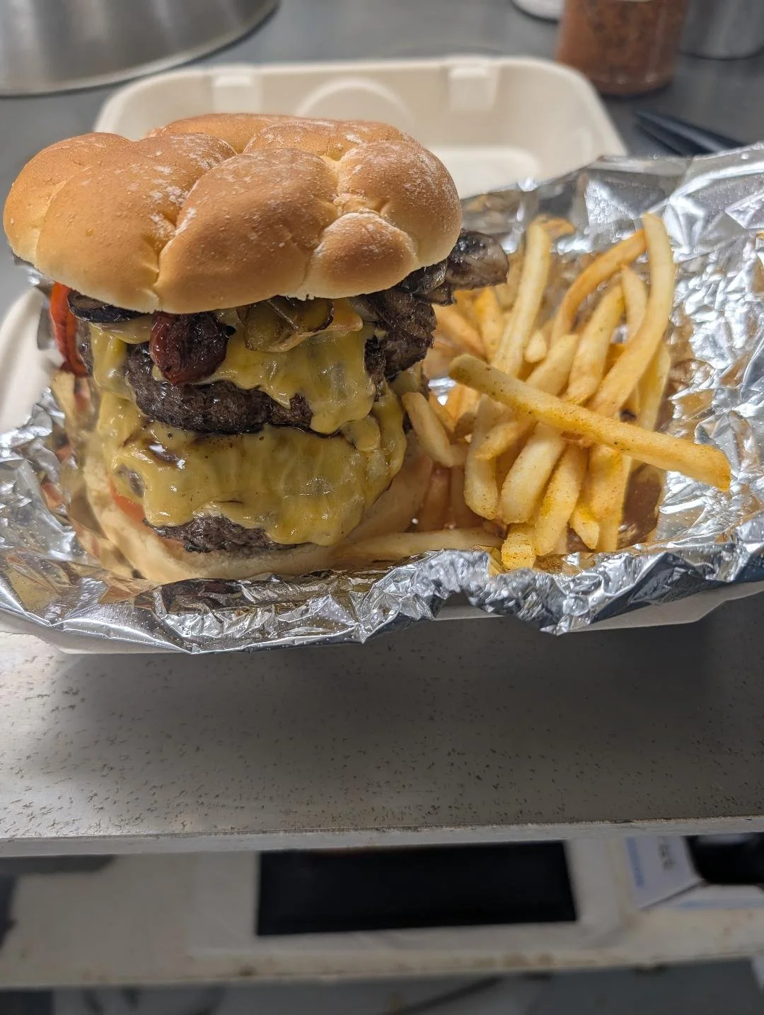 A cheeseburger with multiple beef patties, cheese, grilled mushrooms, and tomato slices, served with a side of French fries in foil.