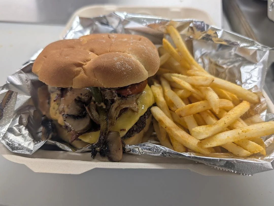 Cheeseburger with sautéed mushrooms and onions, served with French fries in a cardboard tray lined with aluminum foil.