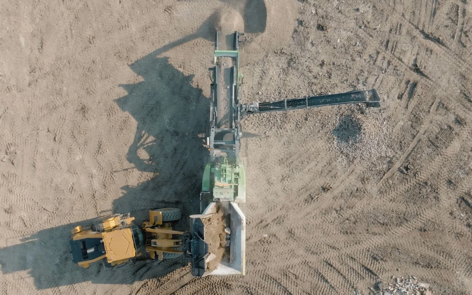 From an overhead view, a construction site showcases a backhoe loader with a yellow arm and black tires, alongside a large piece of machinery with a long black arm, on sandy ground with visible tire tracks and disturbed soil.