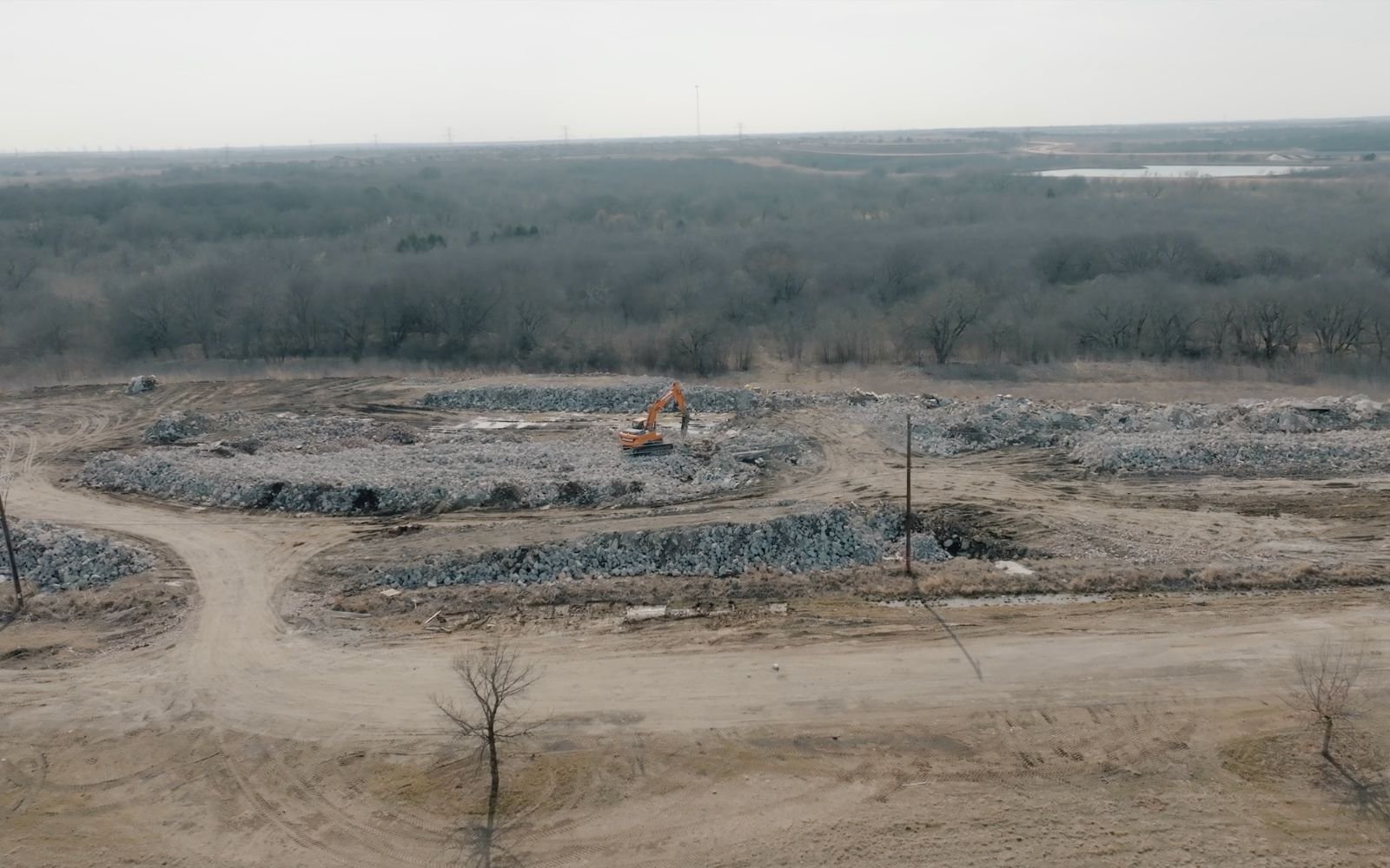 View of a construction site with an excavator operating on a mound of dirt and rocks, surrounded by dirt roads and sparse trees, with a forested area and power lines in the distance under an overcast sky.