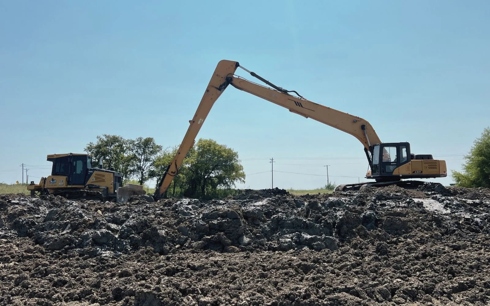 Two construction excavators working on uneven rocky terrain under clear blue sky, with trees and power lines in the background.
