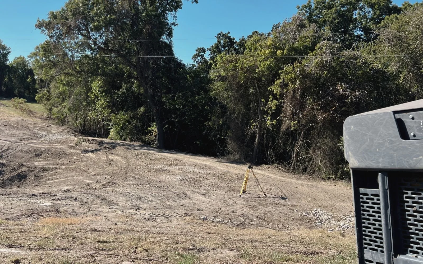 Dirt road with construction equipment, a tripod, and a tree line in the background.