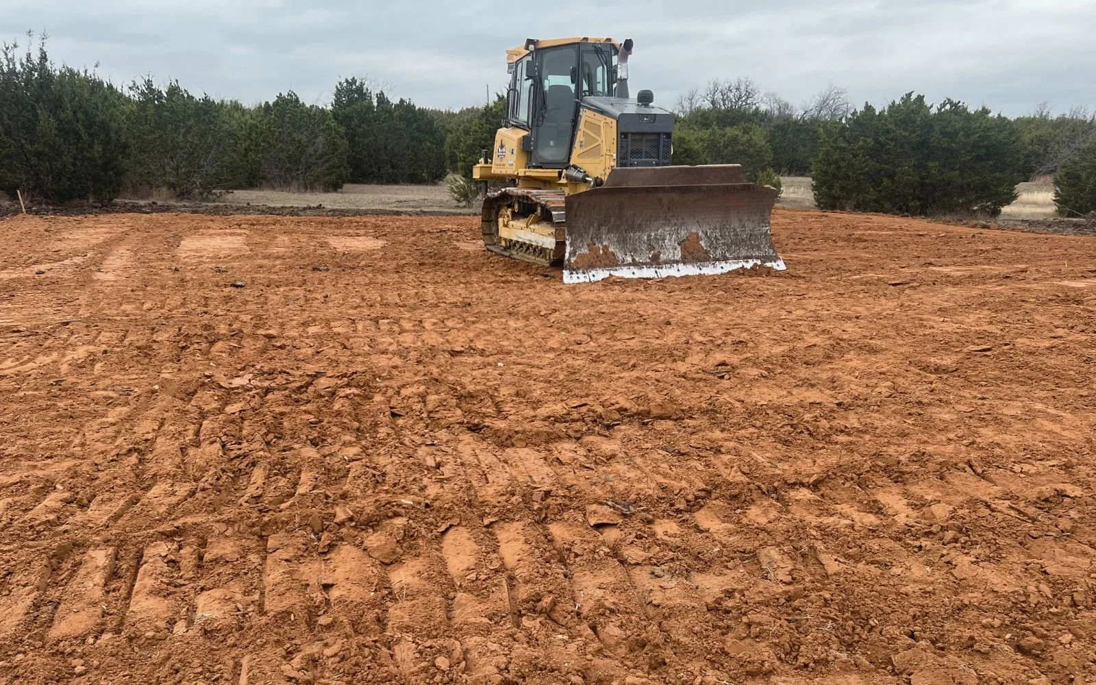 A yellow bulldozer working on a large patch of dirt in an open outdoor area with trees in the background.