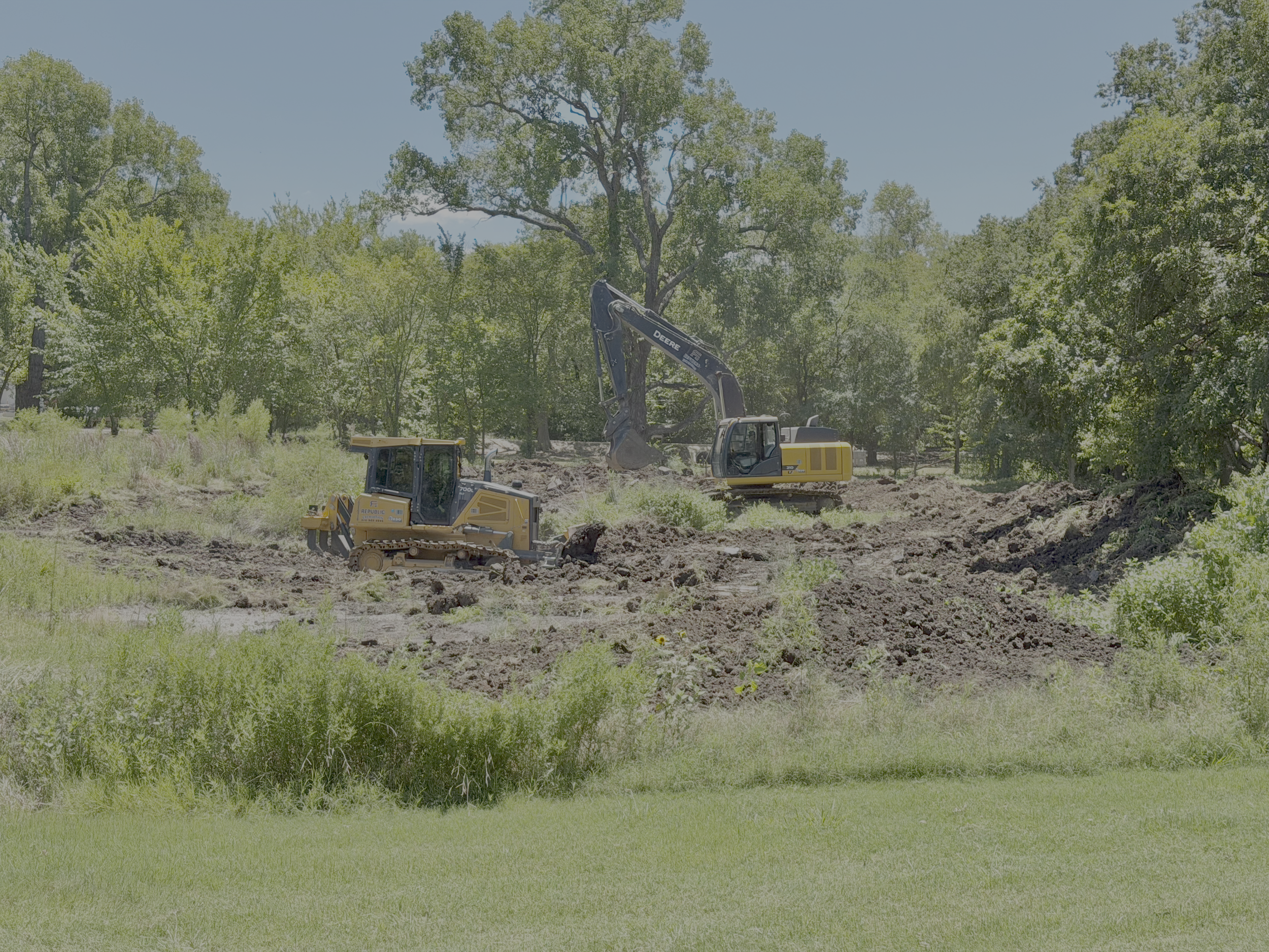Construction excavator digging ground in a grassy area with trees, under a clear blue sky.