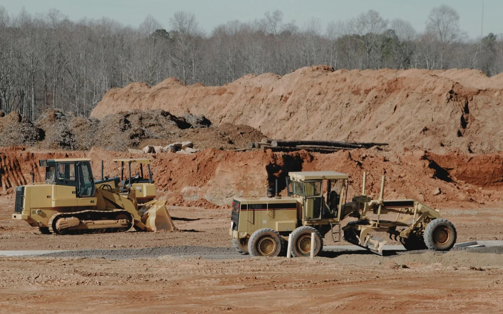 View of a construction site with two heavy machinery vehicles, a bulldozer and a road roller, working on a dirt and gravel surface, with a backdrop of excavated earth and leafless trees in the background.