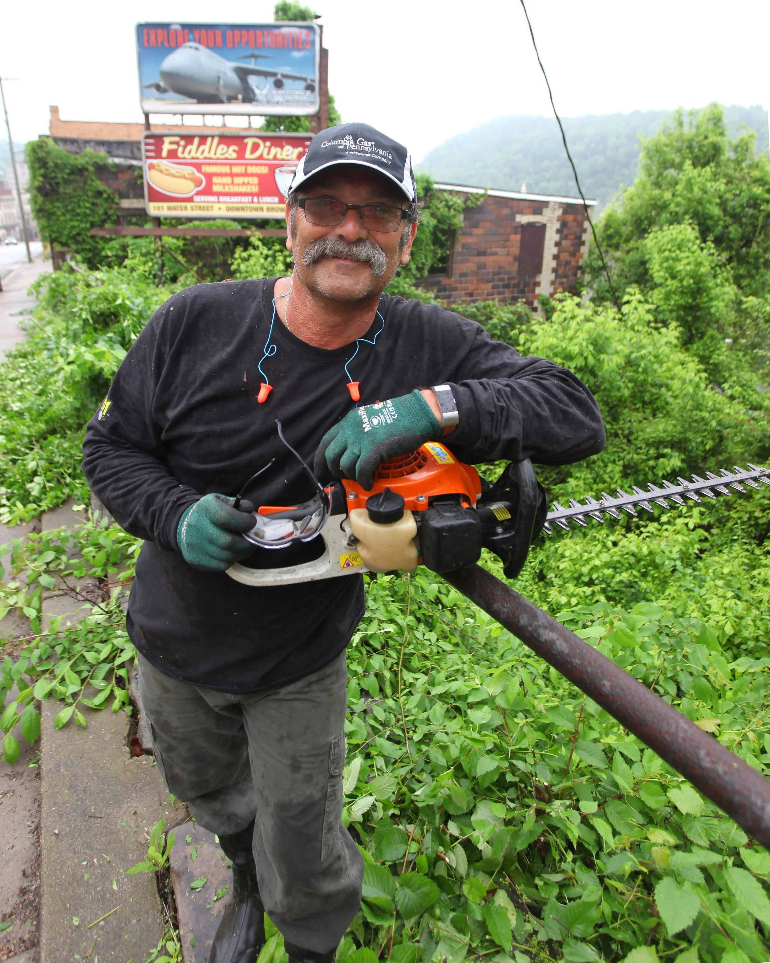 A man with a gray mustache and glasses smiling while holding a leaf blower. He is wearing a black long-sleeve shirt, gray pants, green gloves, a cap that reads 'Columbia Gas of Pennsylvania', and earphones. Behind him are lush green plants and a sidewalk. There are signs in the background, one advertising Fiddles Diner and another with an airplane image and the text 'Explore Your Opportunities'. Mountain scenery and trees are visible in the background.