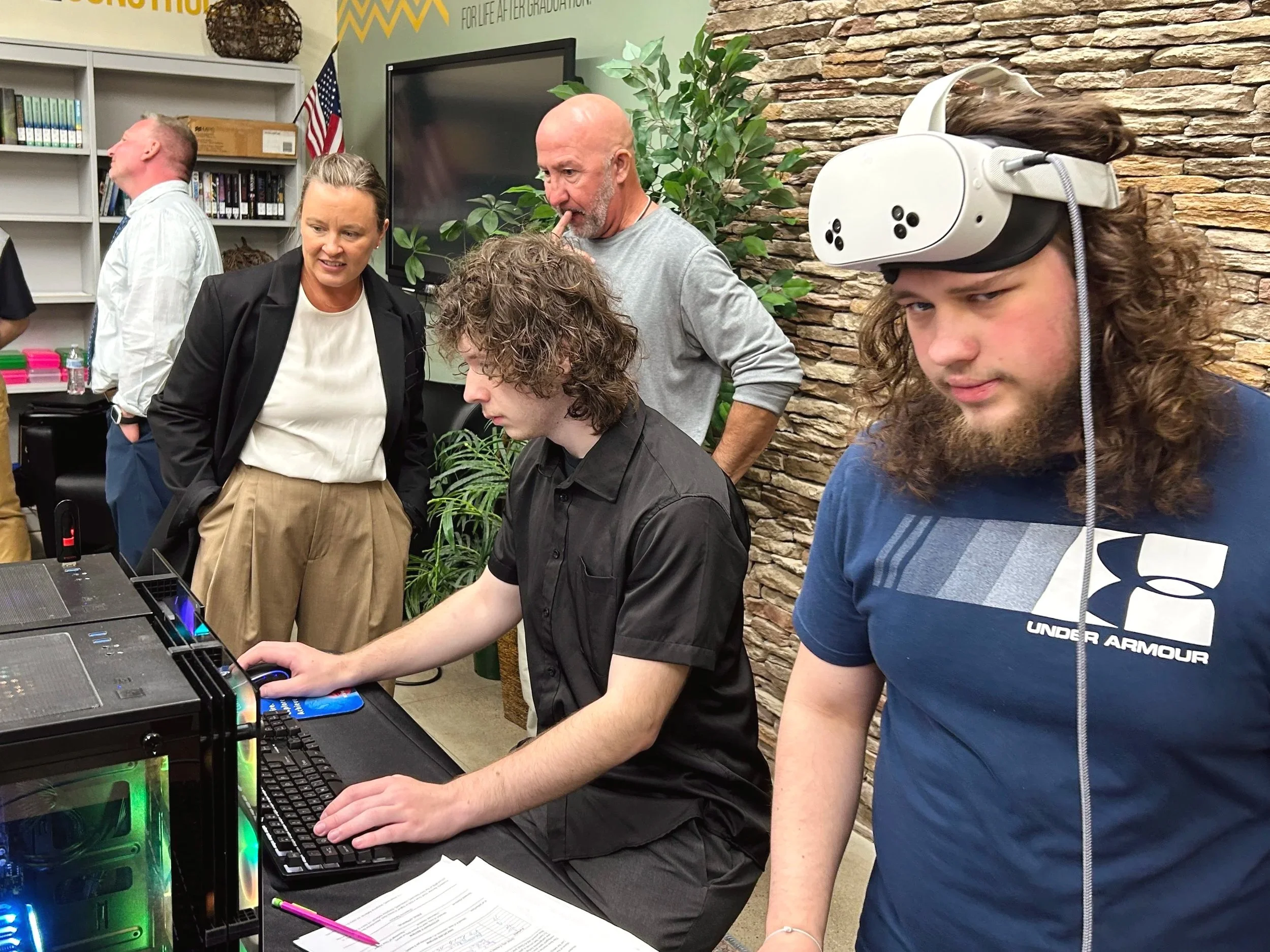 A group of people gathered around a desktop computer in an indoor space with a stone wall and green plants. One person is wearing a virtual reality headset. Others are observing and discussing.